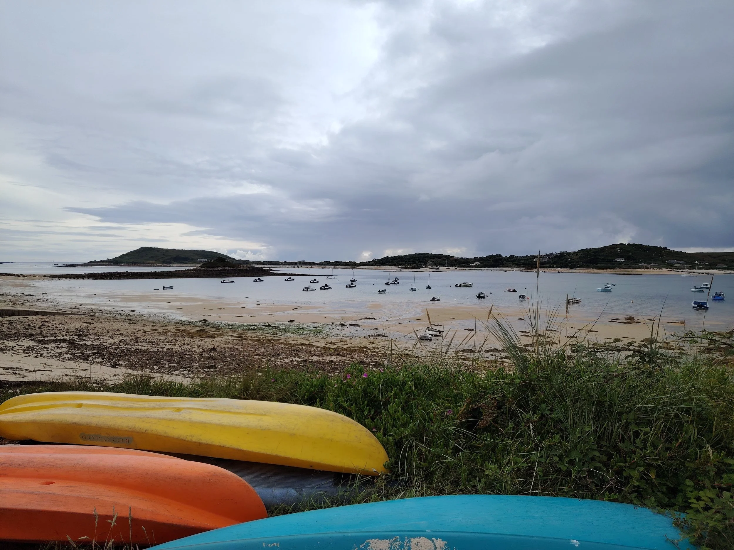 Beach scene with colorful kayaks in the foreground, sandy shore, boats on calm water, and hilly islands under cloudy sky.