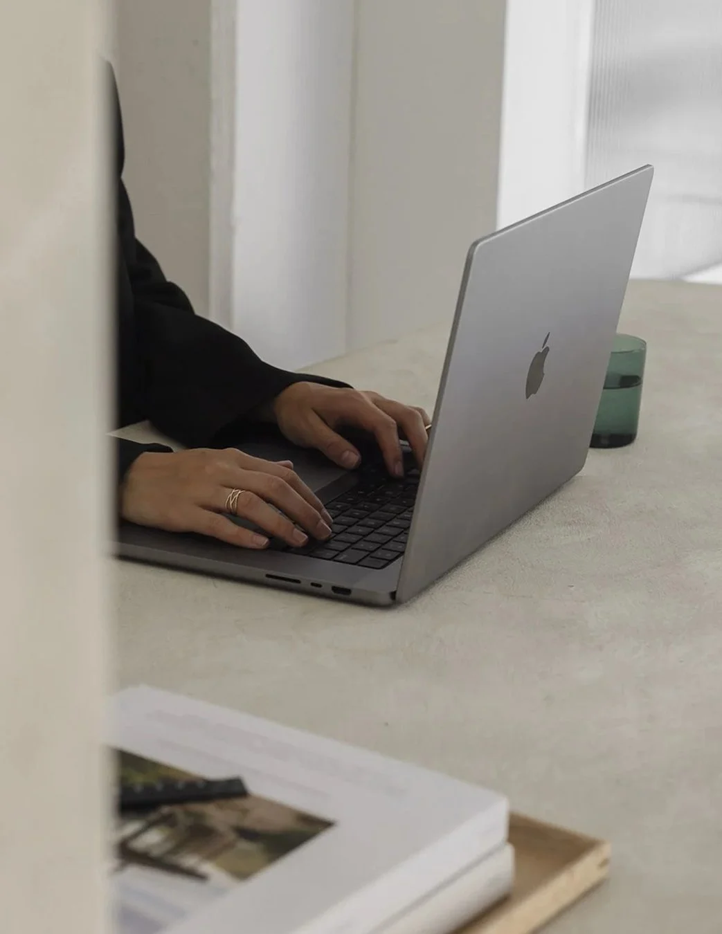 Person wearing black sleeve working on a silver MacBook laptop on a light-colored table, with a book and a small green device nearby.