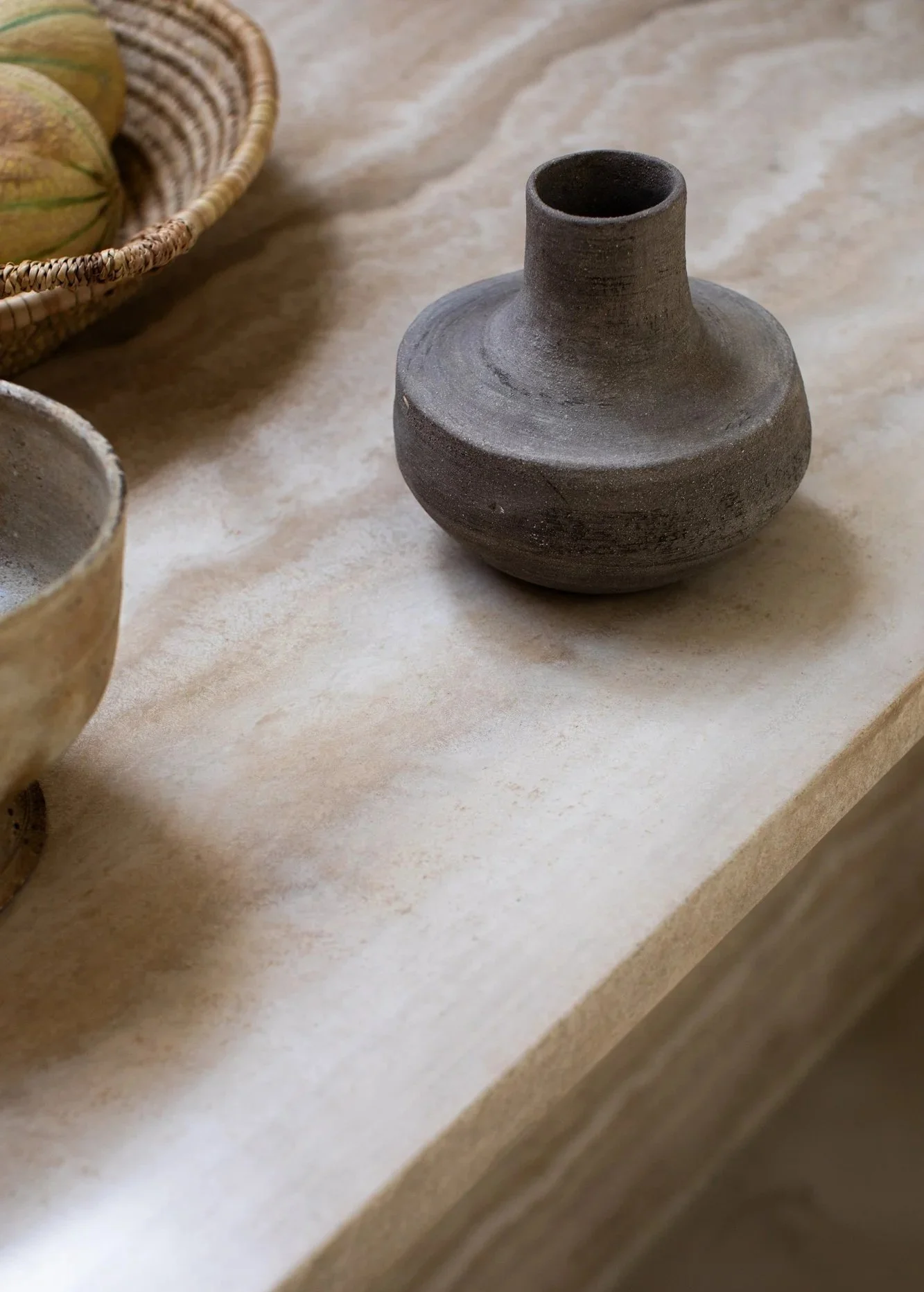 A gray ceramic vase with a narrow opening placed on a light marble surface, with part of a basket with round objects and a bowl visible nearby.