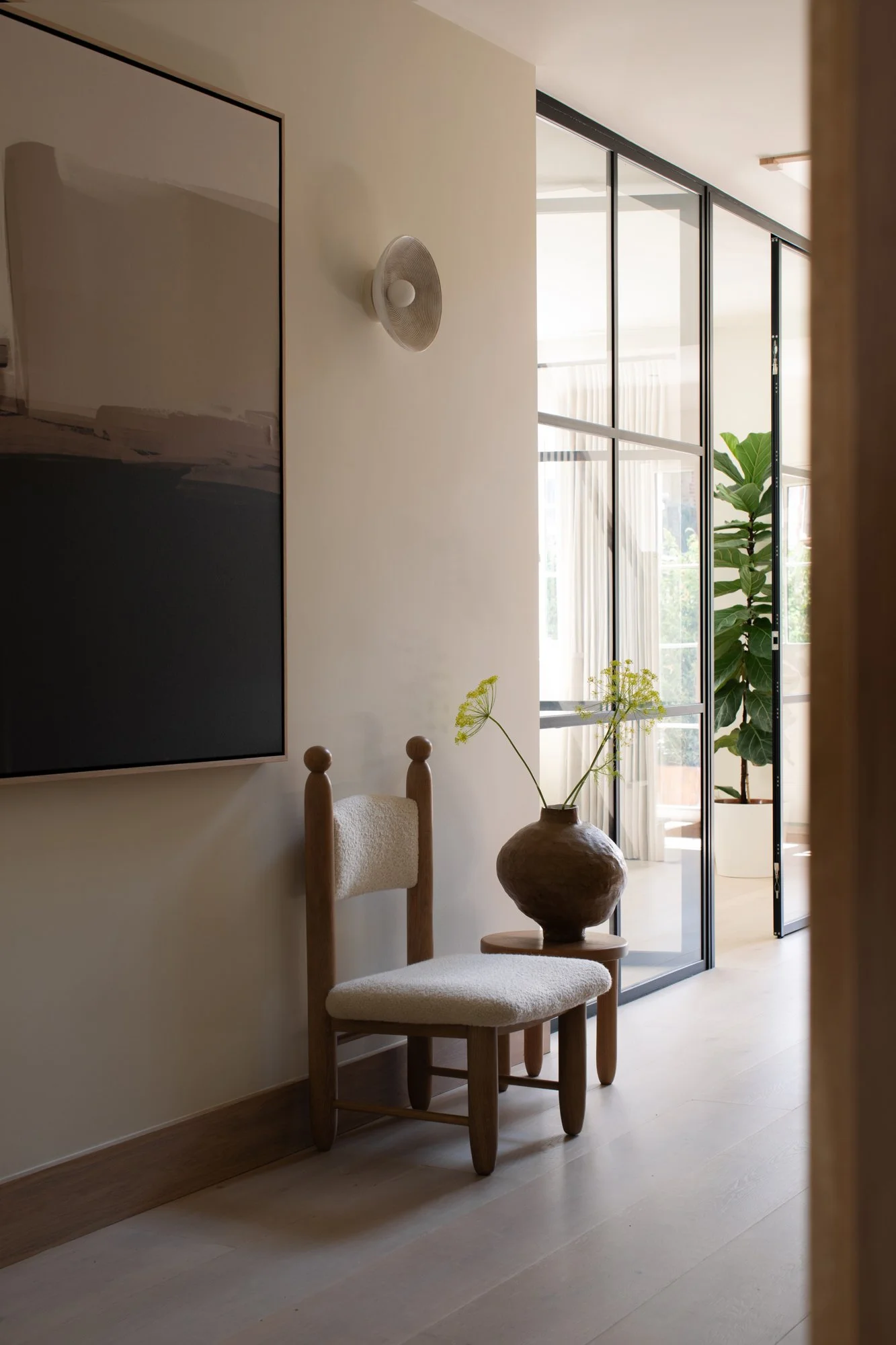 A cozy living space with a wooden chair, a large decorative vase with a plant, large window with sheer curtains, and a wall-mounted light fixture.