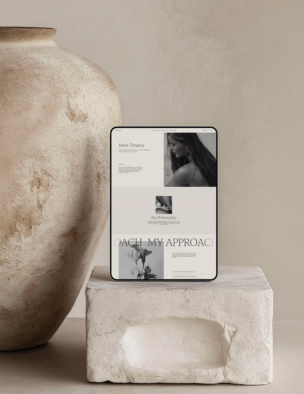 A digital tablet displaying a website with black and white photos of a woman and text, placed on a light-colored concrete block, next to a large textured ceramic vase, against a neutral beige background.