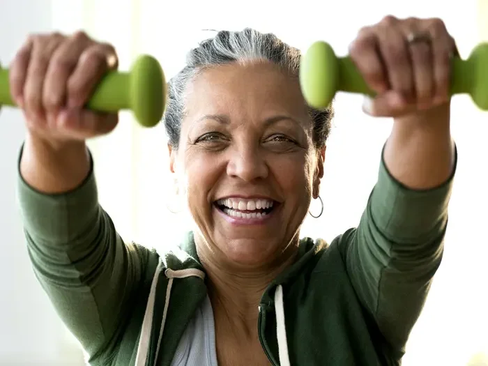 A smiling older woman in a green hoodie exercises with small green dumbbells, looking energized.