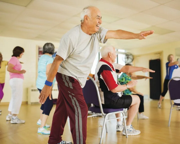 Older adults participating in a seated exercise class, doing arm stretches in a community room.