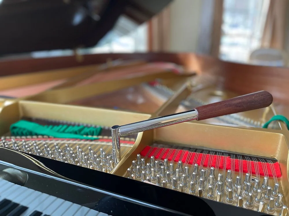 Close-up of the interior of a piano, showing tuning pins, strings, and tuning tools, with a wooden-handled tuning hammer resting inside the piano.