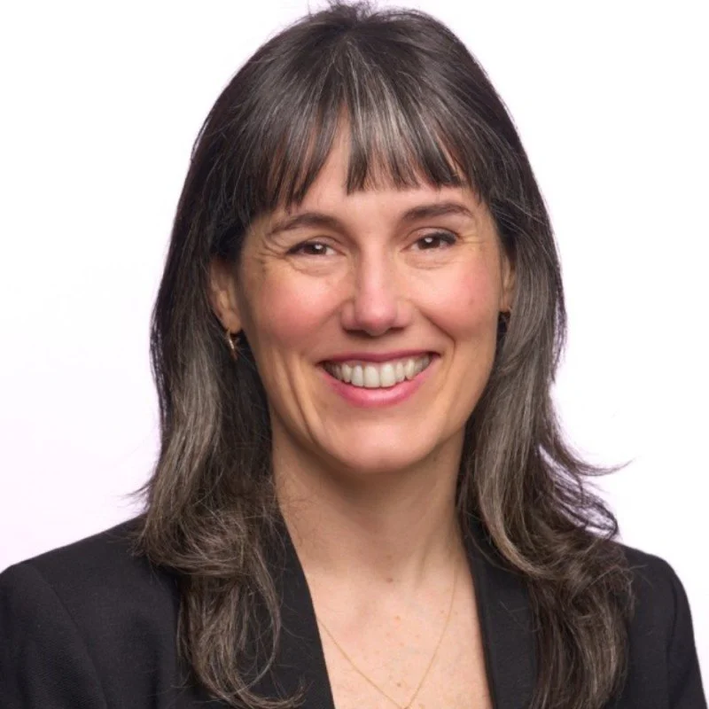 A woman with shoulder-length brown hair and bangs, smiling, wearing a black blazer.
