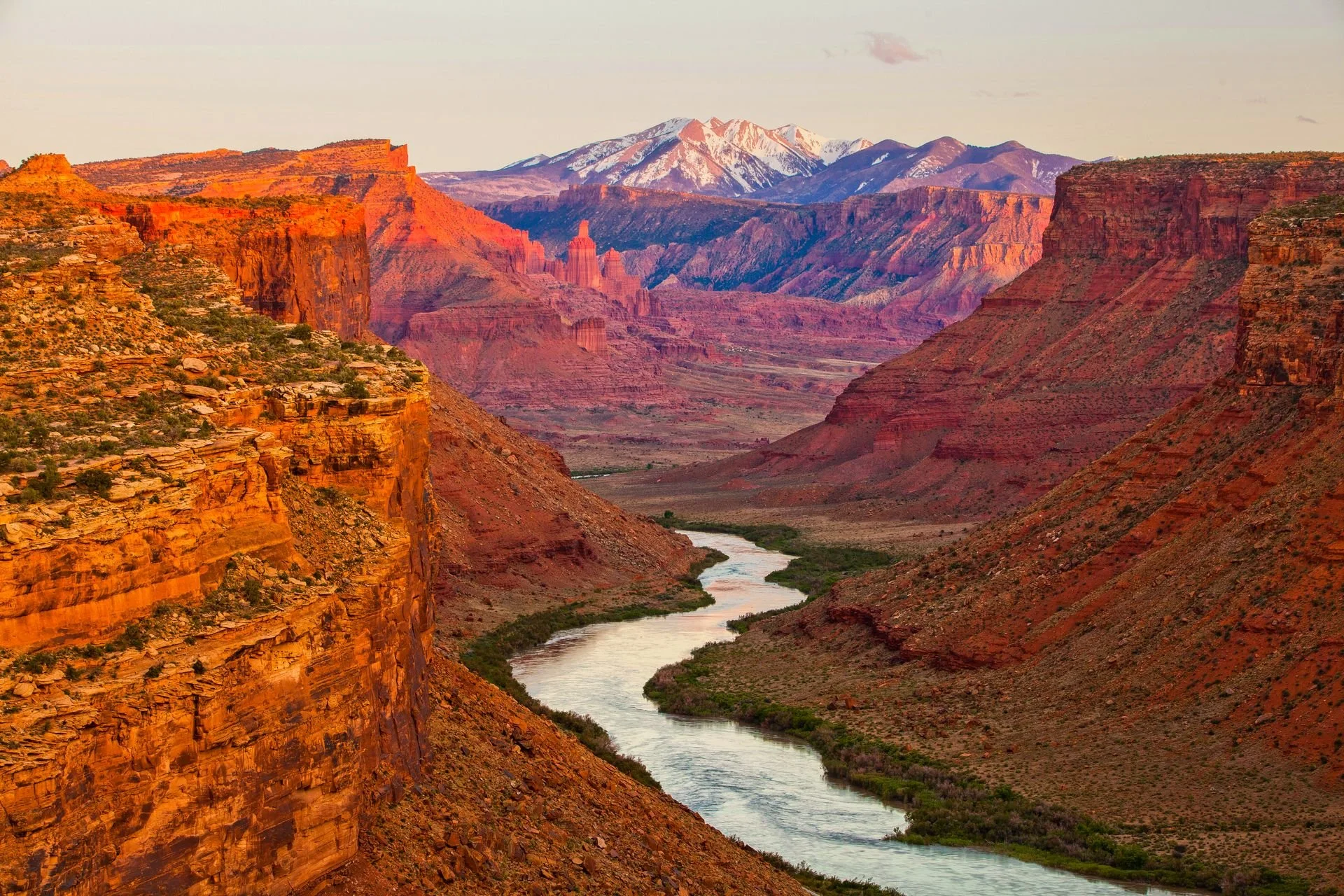 Canyon with red and orange cliffs, a winding river at the canyon floor, and snow-capped mountains in the distance.