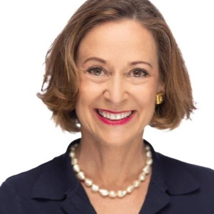 Smiling woman with short brown hair, wearing a dark blazer, pearl necklace, and gold earrings.