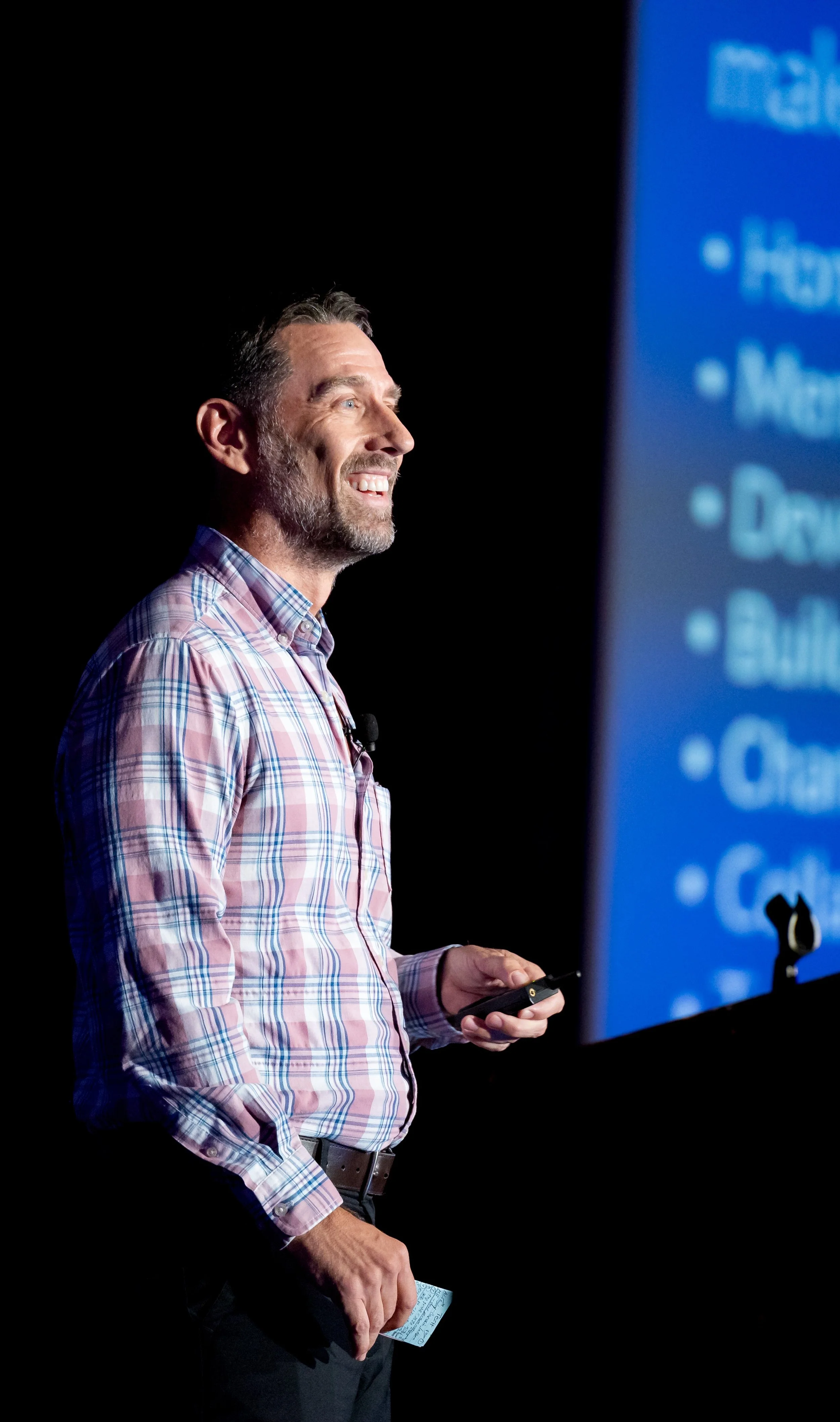 A man (Mike R. Sweeney) with a beard and mustache wearing a pink, blue, and white checkered shirt, standing on a stage, smiling and holding a remote control in his right hand, with a large blue presentation slide partially visible in the background.