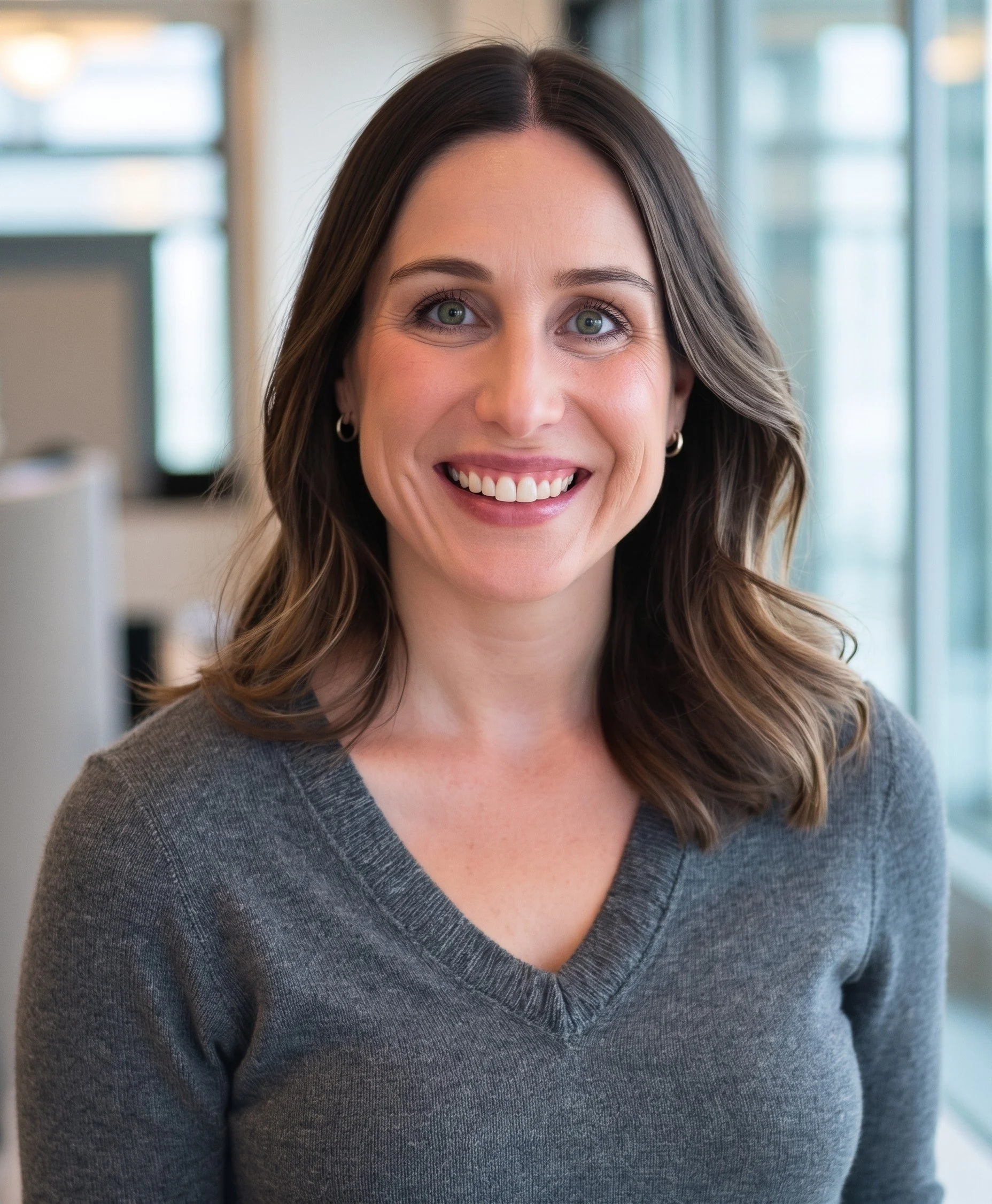 Close-up of a smiling woman (Sydney Cannon) with long brown hair, green eyes, wearing a gray V-neck sweater, standing in a modern office environment with glass windows.