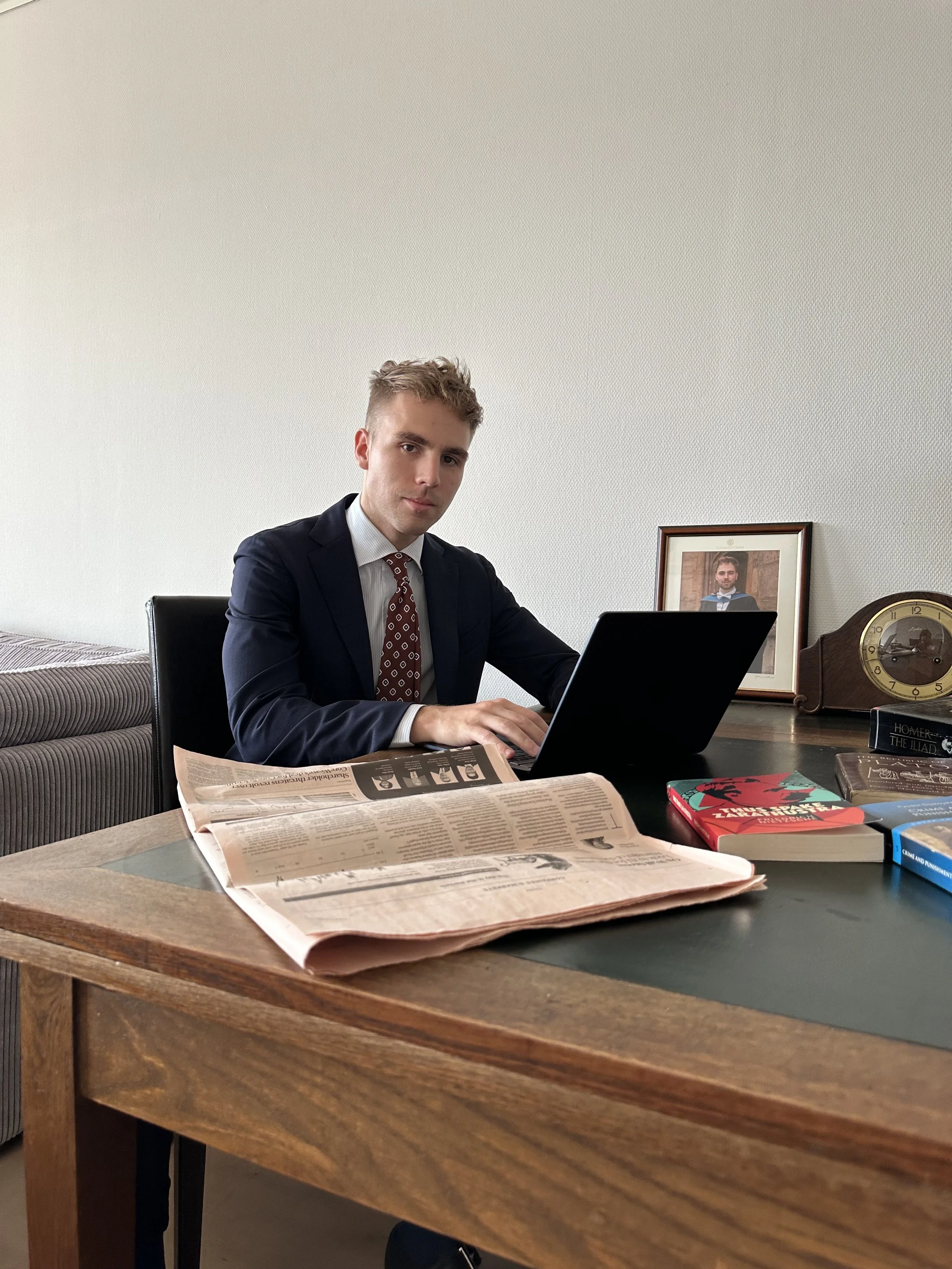 A young man in a suit sitting at a wooden desk working on a laptop with newspapers and books, in an office with a framed photo and a clock in the background.