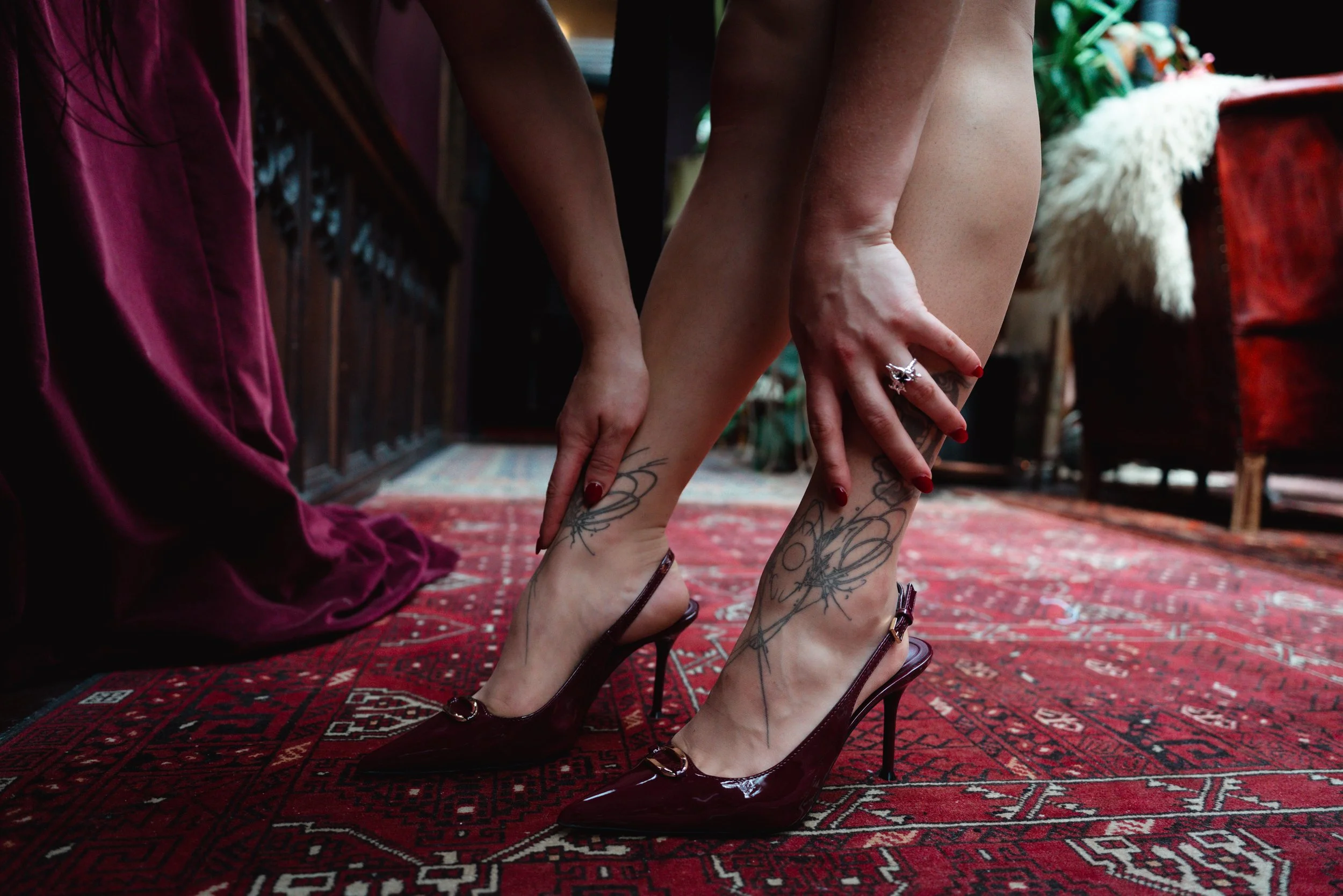 A woman wearing burgundy high heels is adjusting her tattoos on her legs, which are visible as she is partially unclothed. She is standing on a patterned red rug in a cozy room with wooden furniture and plants.