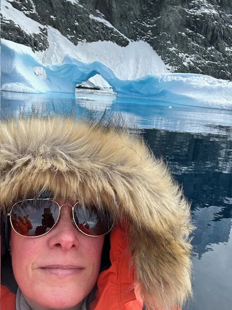 A person wearing sunglasses and a hooded jacket with fur trim in front of an icy glacier and calm water in a cold, polar environment.