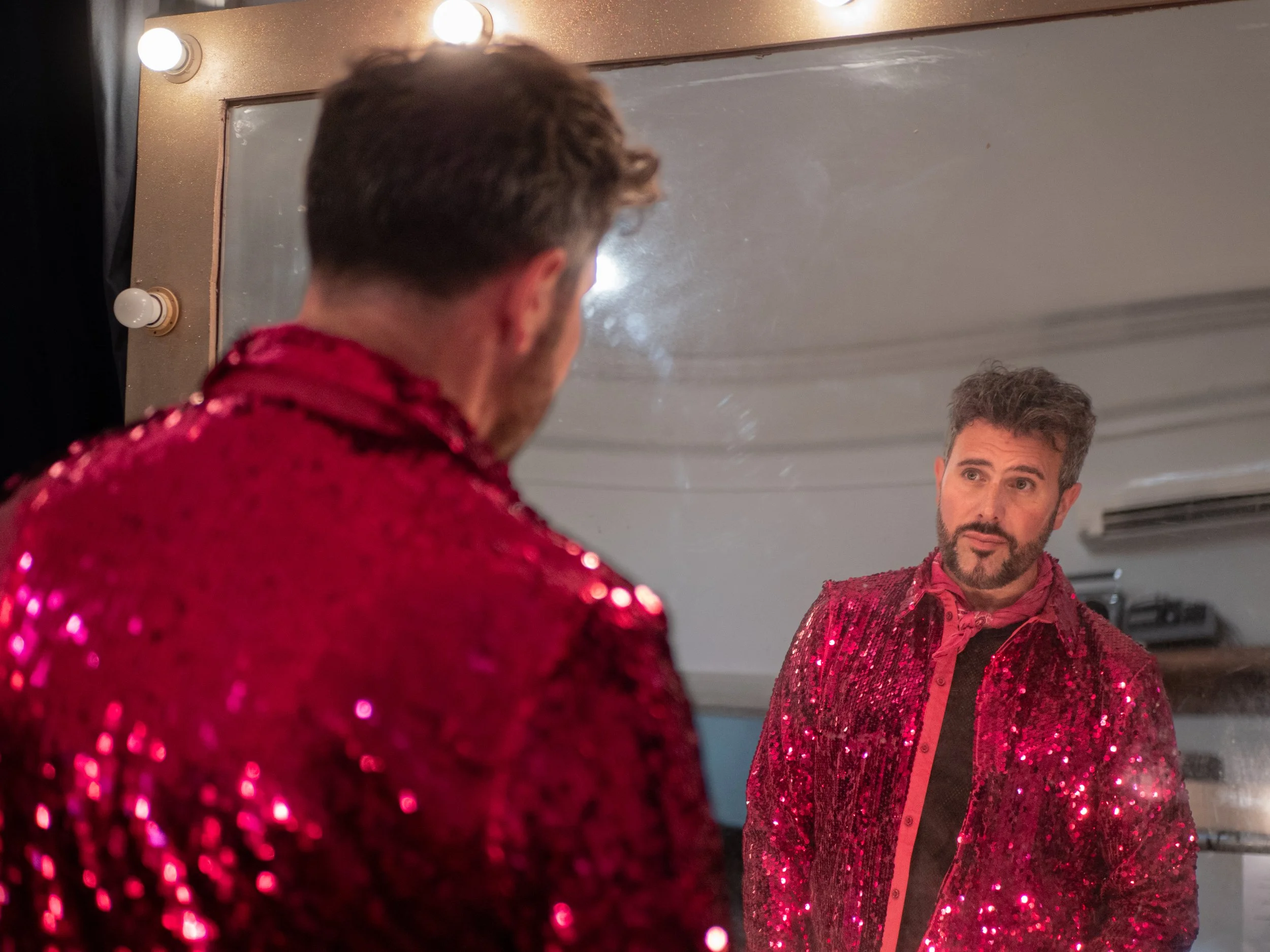 A man looking at himself in a mirror, wearing a shiny red sequined jacket, with a pink bandana around his neck, and green eyes with a beard. He is preparing for a show.
