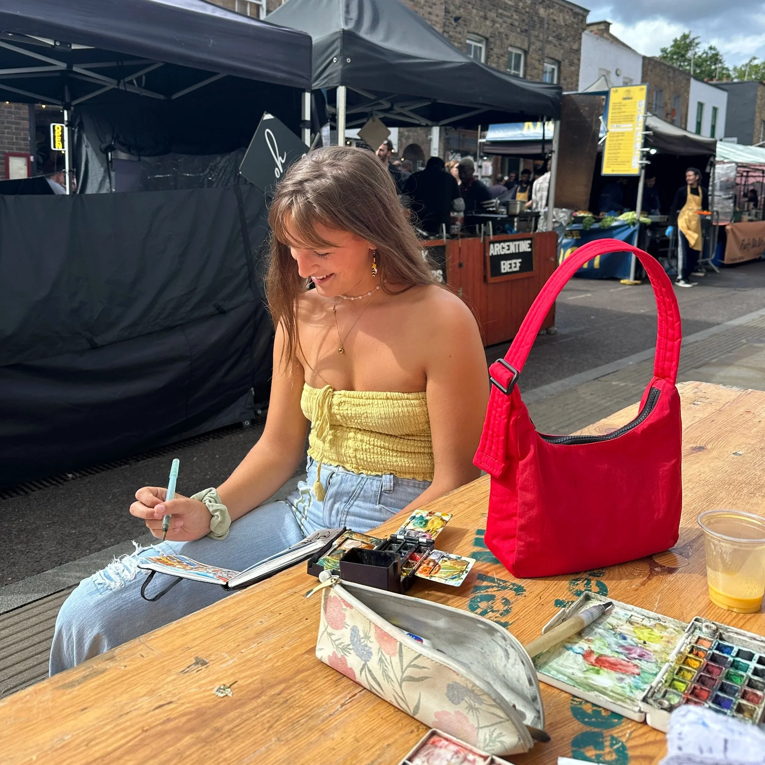 A woman sitting at a wooden table with art supplies, smiling and writing in a sketchbook. She has long brown hair, wearing a yellow tube top, light blue jeans, and multiple jewelry pieces. There is a red bag on the table, along with watercolor paints, brushes, and a cup of yellow liquid. The background shows an outdoor market with tents and people.