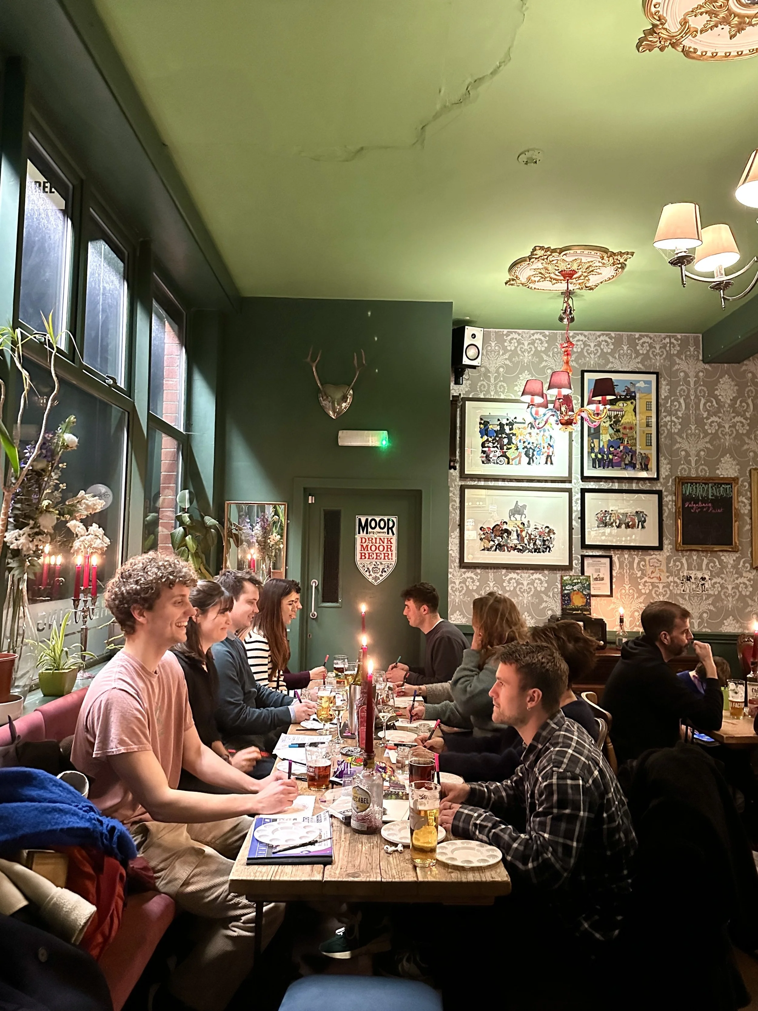 A group of people sitting around a long wooden table in a cozy restaurant, celebrating with drinks and candles, with decorative artwork and plants in the background.