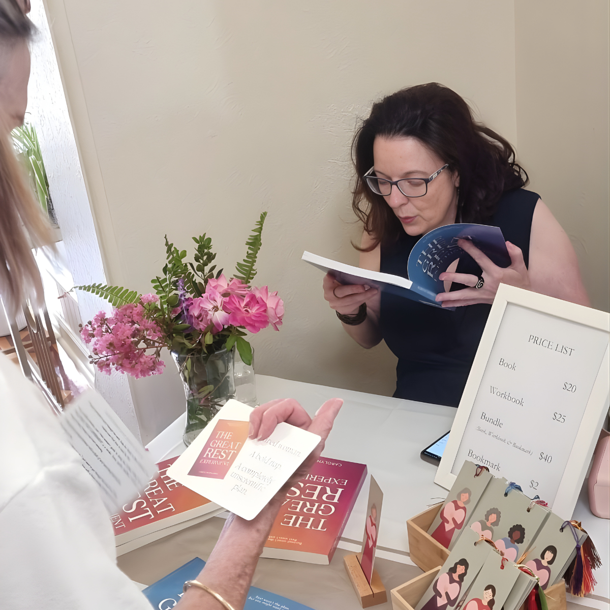 A woman with black hair and glasses behind a table at a bookstore, looking at a book, with a sign listing prices of various items on the table.