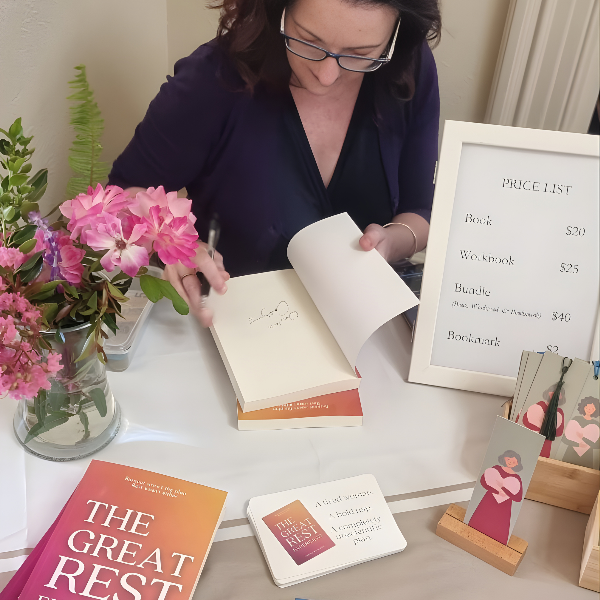 A woman with dark hair and glasses signing a book at a table with a sign displaying prices for books, workbooks, bundles, and bookmarks. The table is decorated with pink flowers in a glass vase, and there are promotional materials including a book titled 'The Great Rest' and a small display card with an illustration of a woman.
