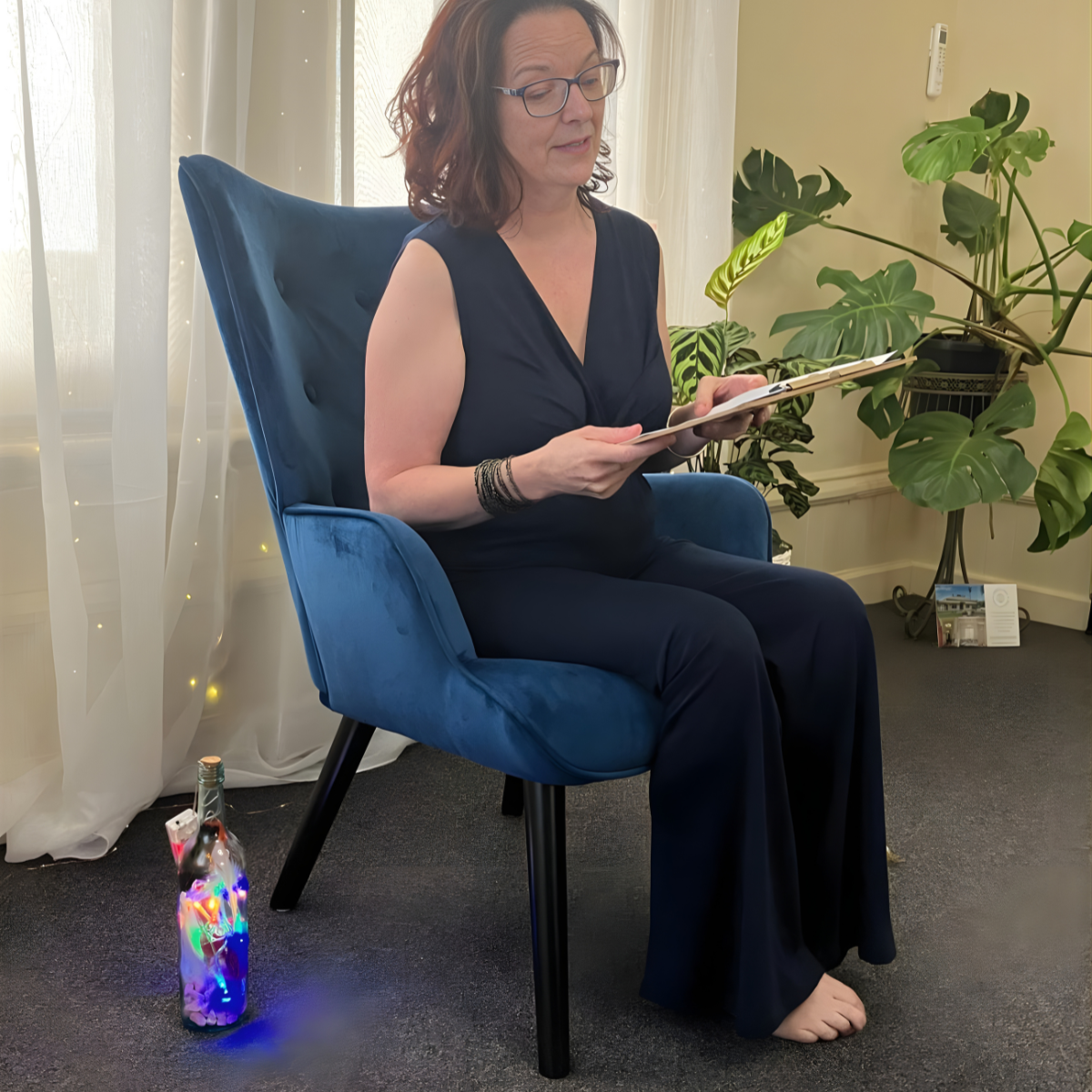 A woman with glasses sitting in a blue upholstered chair, reading a folder or magazine. She is in a bright room with curtain-covered windows and green houseplants. There is a bottle with colorful LED lights on the floor beside her.