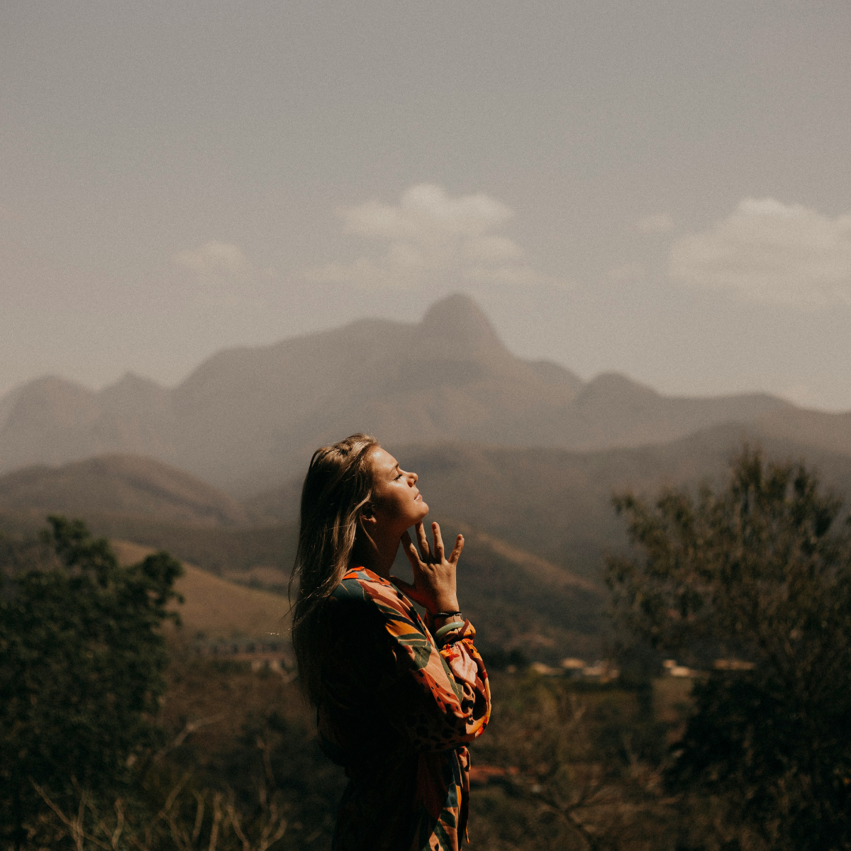 Spiritual rest - A woman with long hair, wearing a colorful dress, stands outdoors with her eyes closed and hand near her face in a contemplative pose, with mountains and trees in the background.
