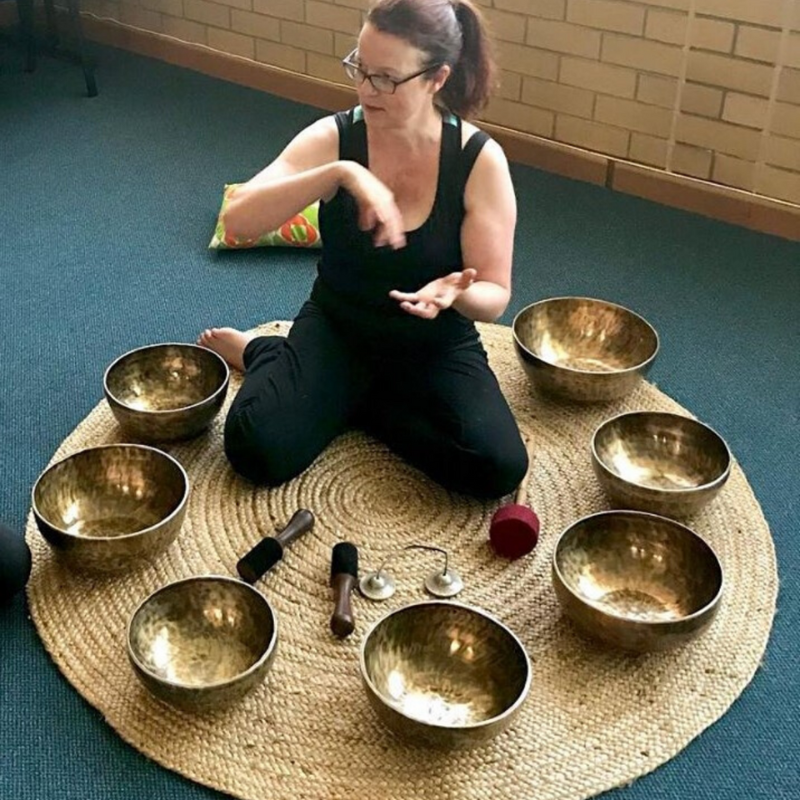 A woman sitting cross-legged on a woven mat, surrounded by eight metallic singing bowls and various percussion instruments, gesturing with her hands.