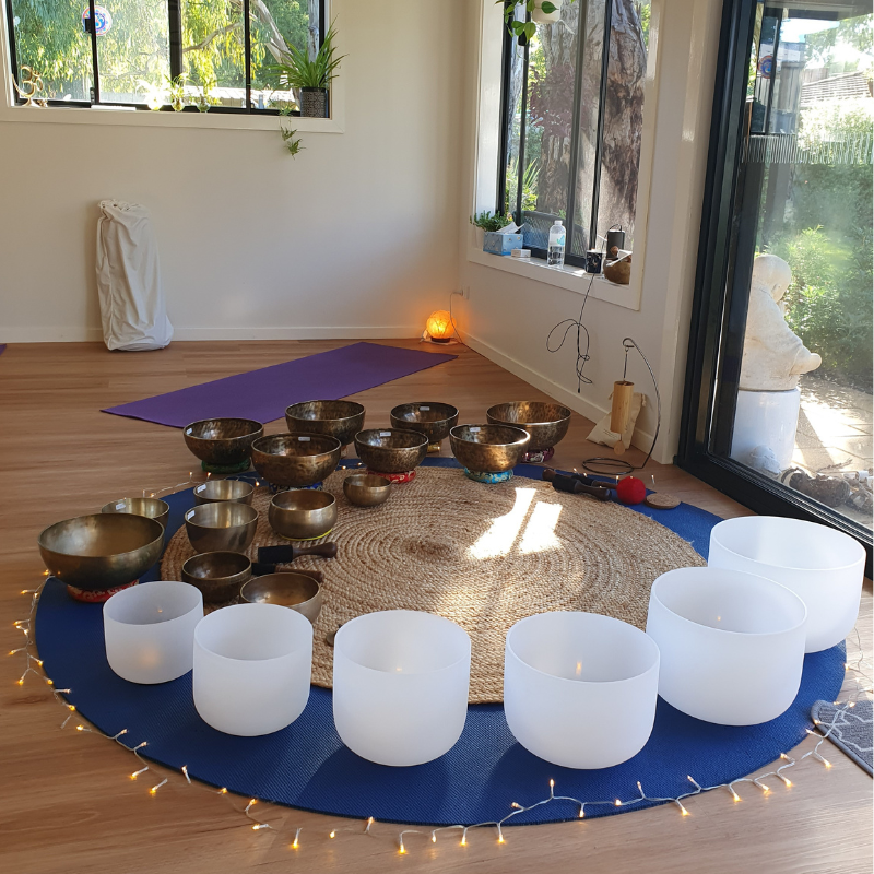 A room set up for sound healing or meditation with crystal singing bowls, white LED candles, a purple yoga mat, and a natural fiber rug on a wooden floor. Window with greenery outside, and sunlight coming in.