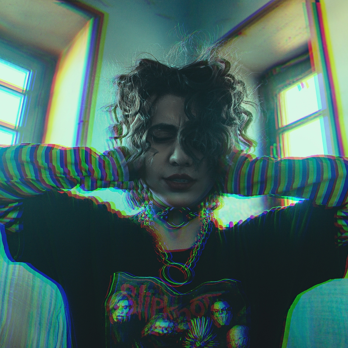 Sensory rest - A person with curly hair, wearing a graphic t-shirt, layered necklaces, and rainbow-striped long sleeves, poses indoors with their hands behind their head, surrounded by colorful, distorted light effects.
