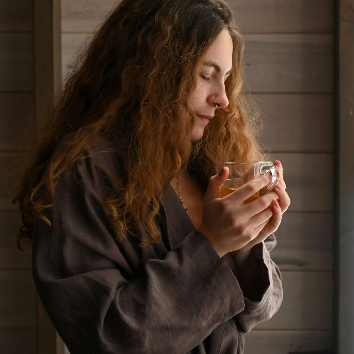 Mental rest - A woman with long curly red hair and a nose ring holds a glass cup of tea with both hands, standing against a wooden background.