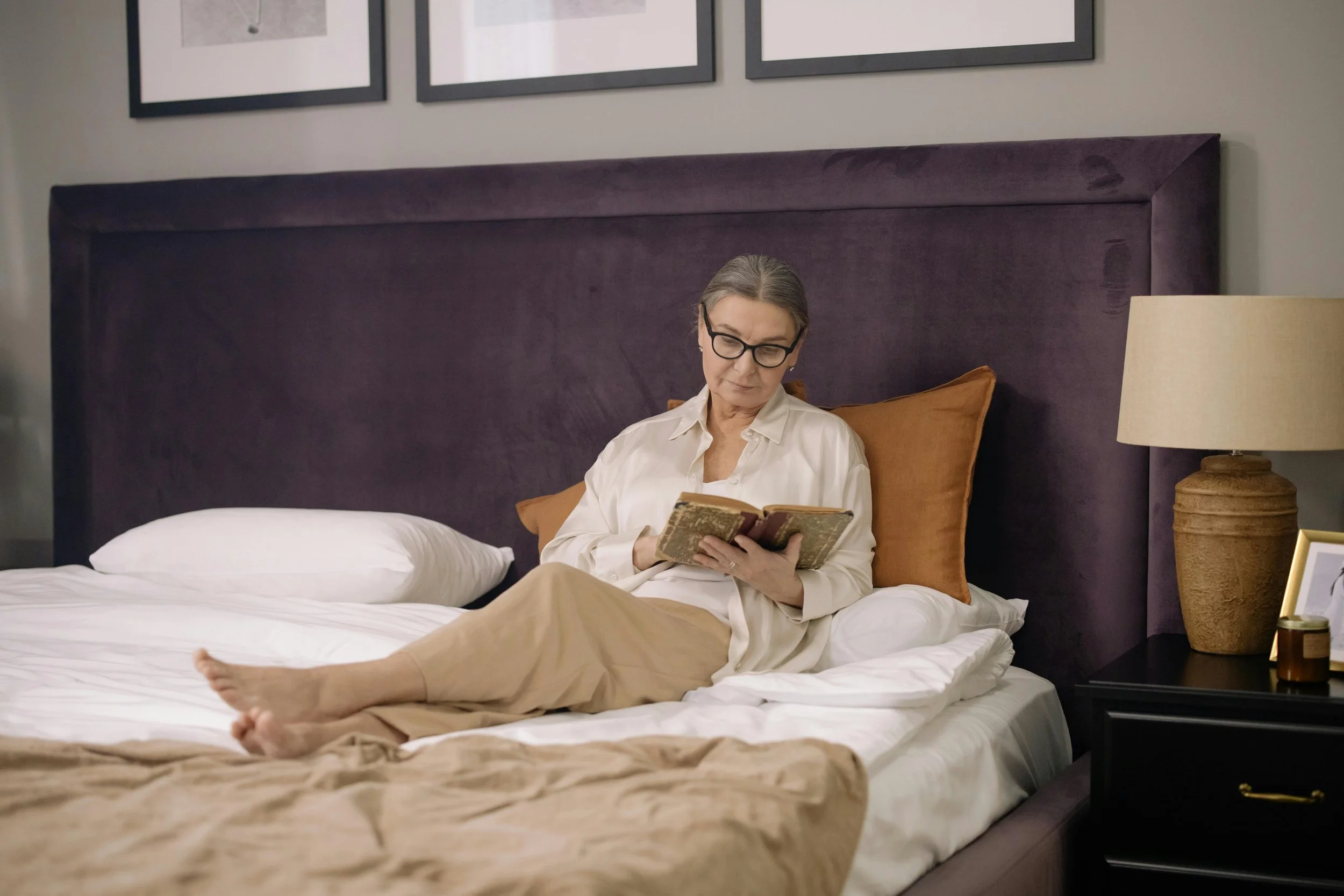 Social rest - An elderly woman with glasses reading a book while sitting on a bed with beige sheets and pillows in a modern bedroom.
