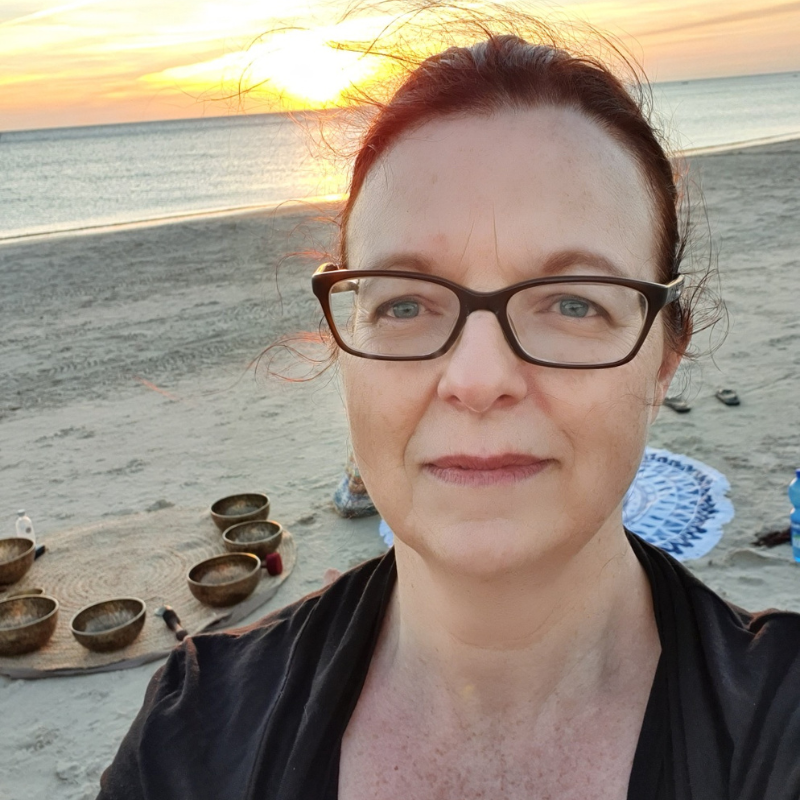 A woman with glasses taking a selfie on the beach during sunset, with singing bowls and a patterned blanket visible in the background.