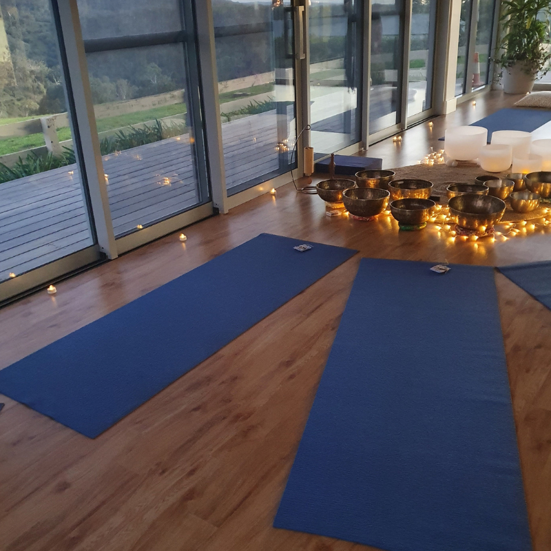 Yoga mats arranged in a circle on wooden floor near large windows, with singing bowls and candles on the side.