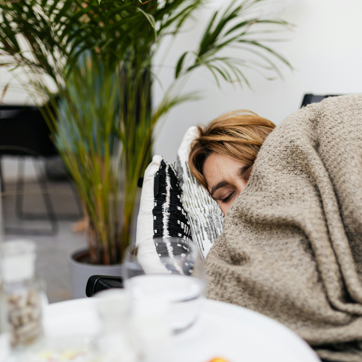 Physical rest - A woman with light brown hair sleeping on a pillow under a cozy blanket at a table, with a large green potted plant in the foreground.