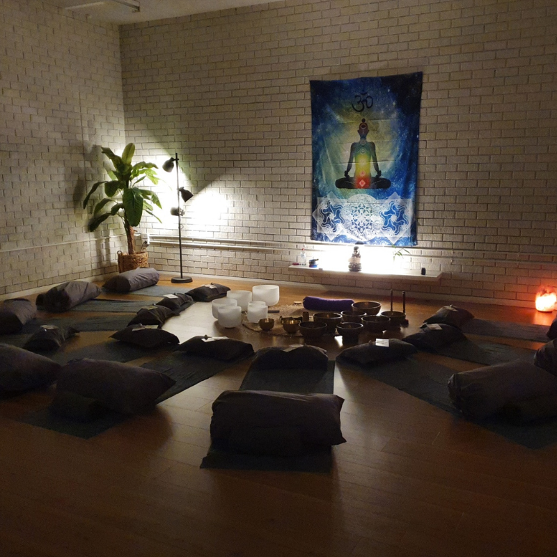 A meditation space with black cushions arranged in a semicircle around plates and bowls, a large plant in a basket, a tall reading lamp, and a colorful tapestry with a meditative figure hanging on a white brick wall, illuminated by soft lighting.