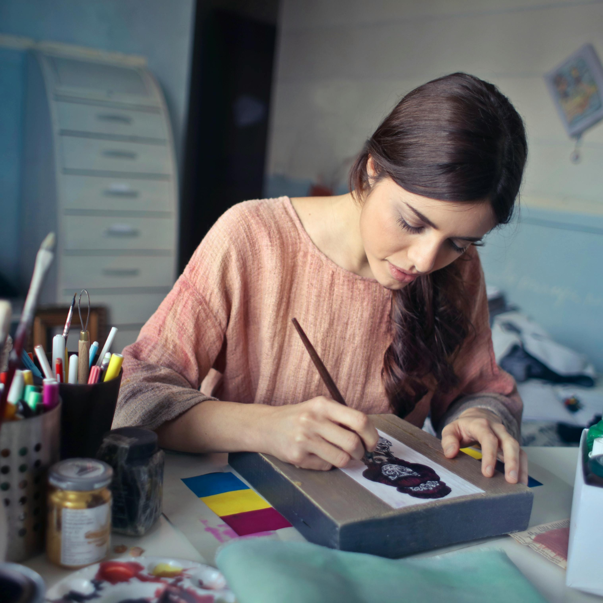 Creative rest - A woman working on a mixed media art piece at a desk with art supplies, including brushes, markers, and paints, in a room with a cluttered background.