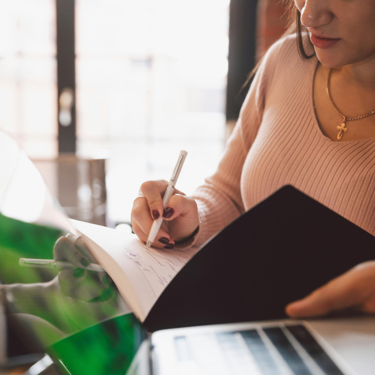 A woman in a pink ribbed top writing in a notebook with a pen, sitting at a desk with a laptop and a soda bottle.