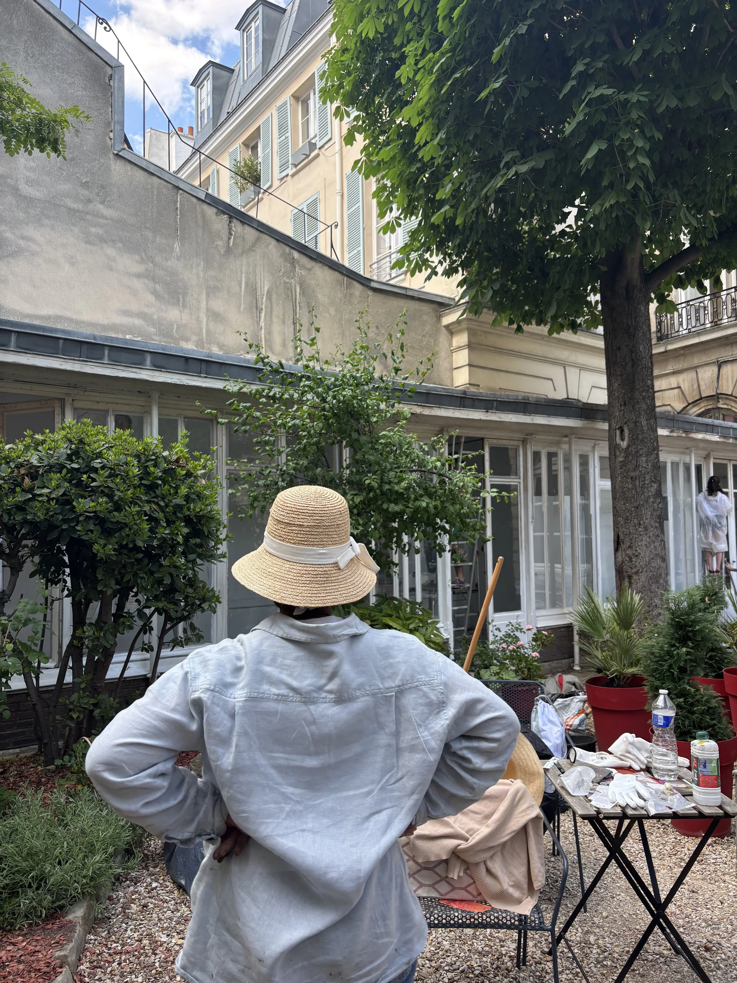 Une personne vue de dos portant un chapeau en paille et une chemise blanche, debout dans un jardin intérieur avec des plantes en pot, des tables, et une grande forêt à proximité. Le fond montre une maison avec des fenêtres en verre et une façade beige, sous un ciel partiellement nuageux.