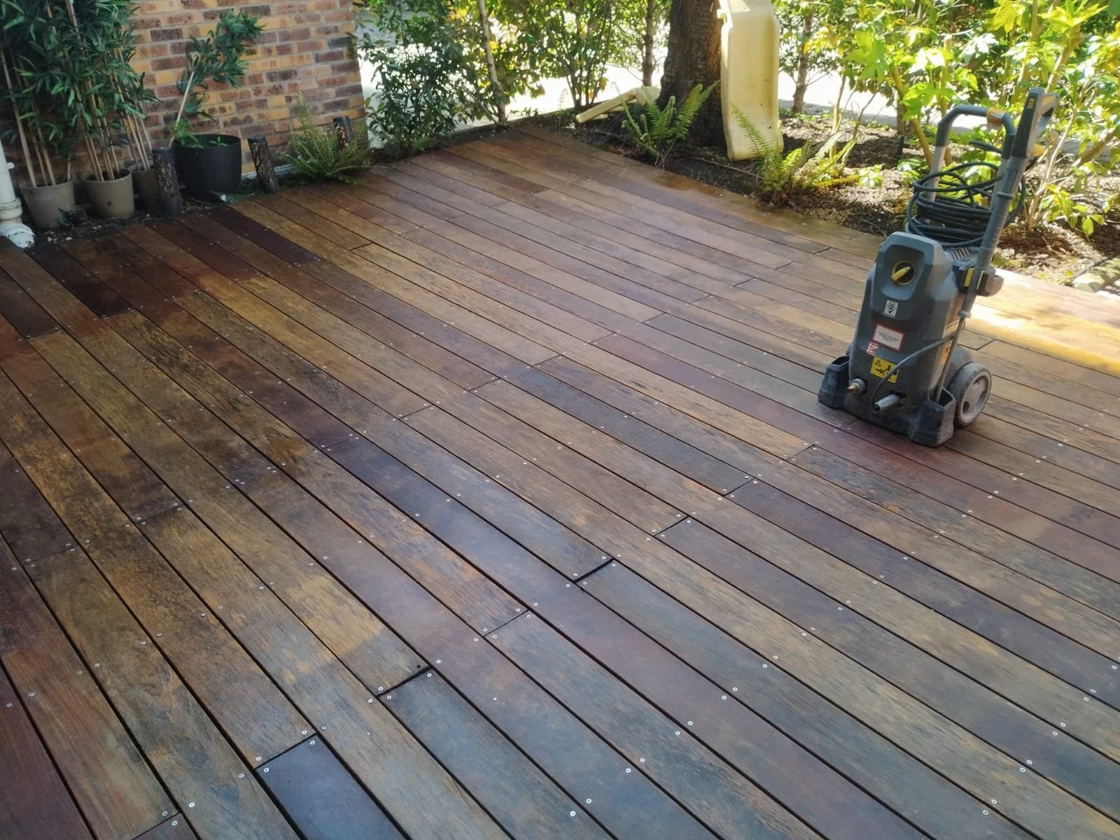 Un patio en bois en cours de nettoyage ou de finition, avec une machine de nettoyage ou de polissage sur le sol. Il y a quelques plantes en pots et des arbustes en arrière-plan.