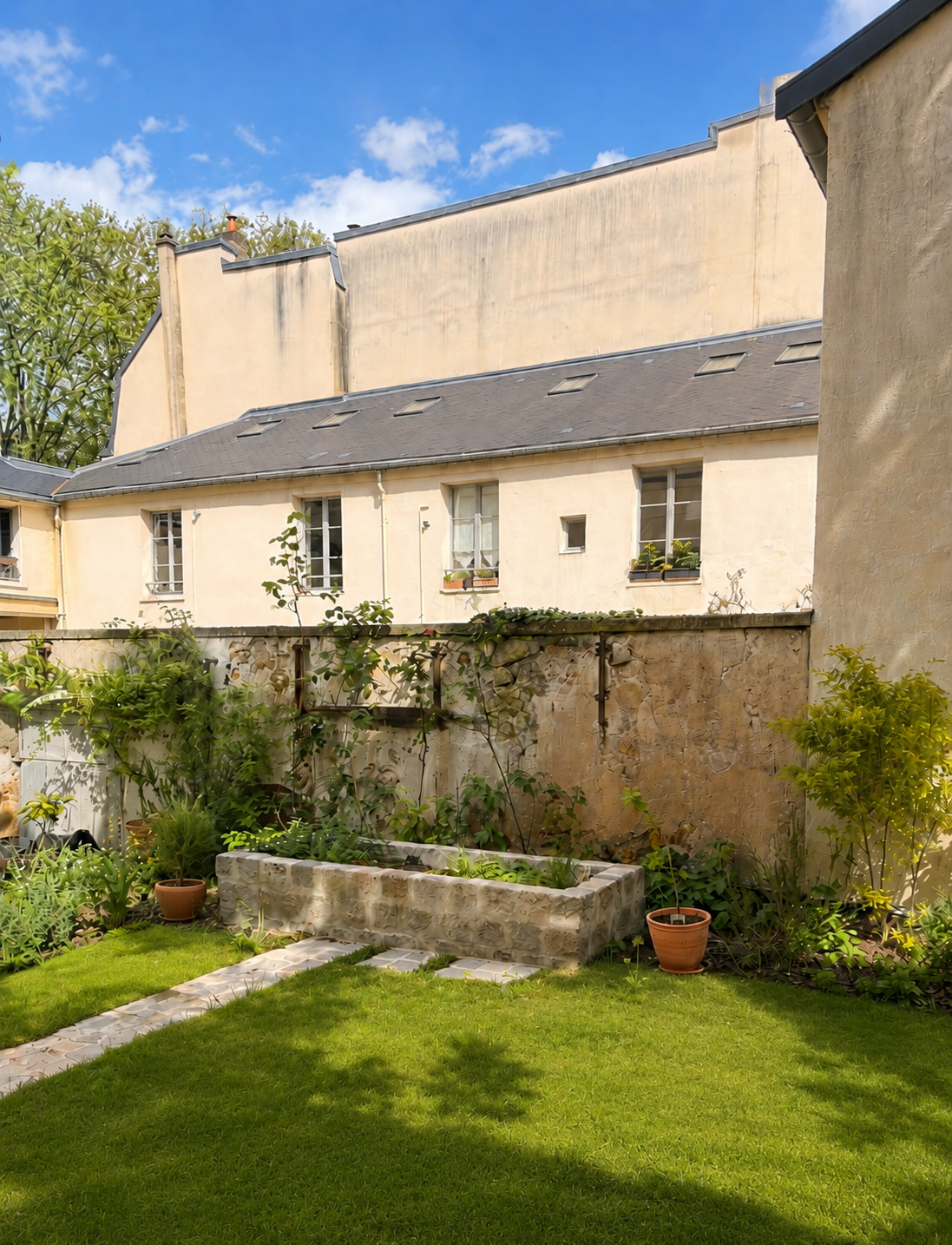 Jardin avec pelouse verte, un planter en pierre contenant des plantes, plusieurs pots de fleurs, mur en pierre et maison en arrière-plan sous un ciel bleu avec quelques nuages.