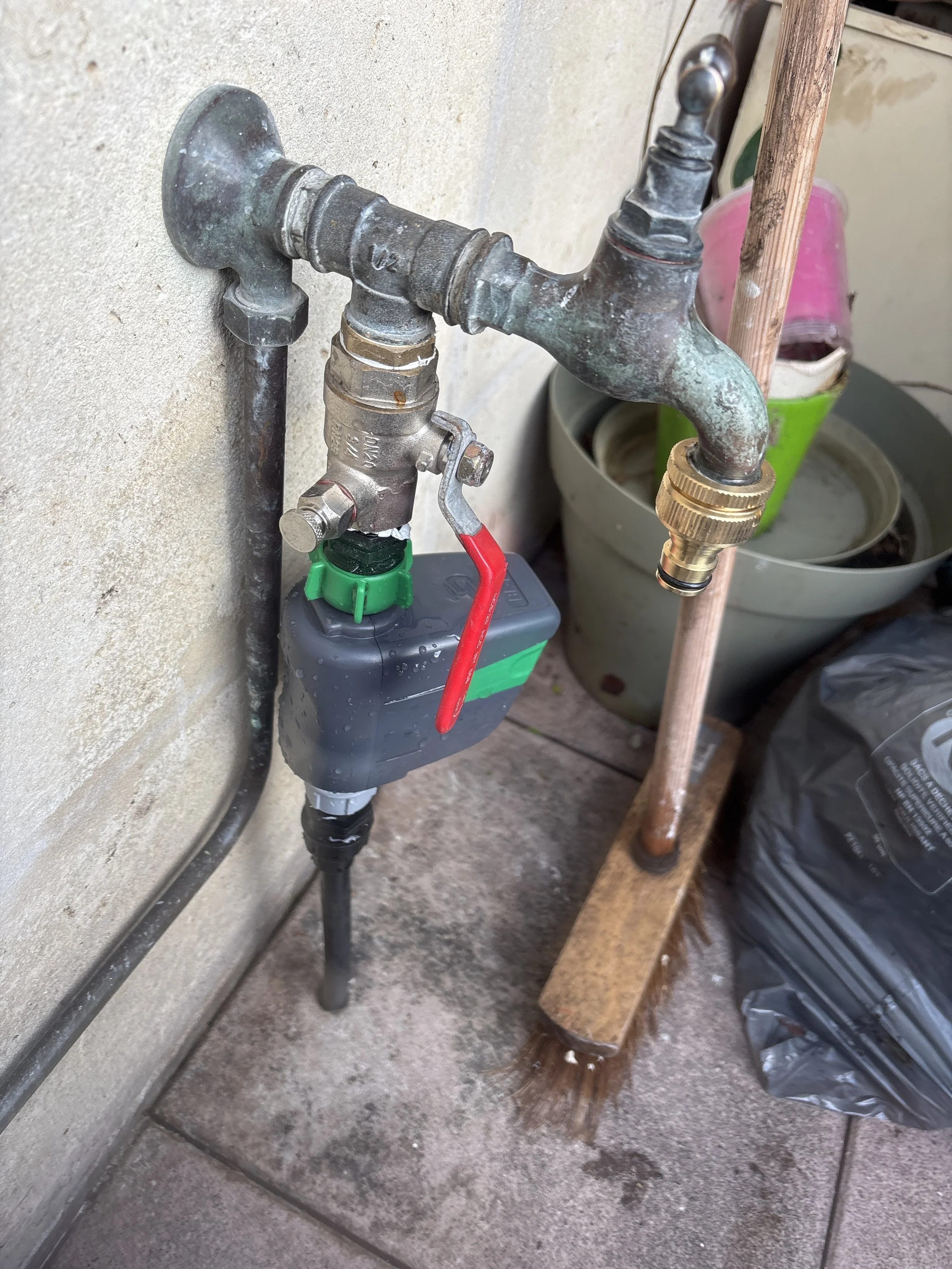 Robinet d'eau extérieur avec un dispositif de contrôle et un tuyau attaché, situé sur un mur près du sol, avec un seau et des outils à proximité.