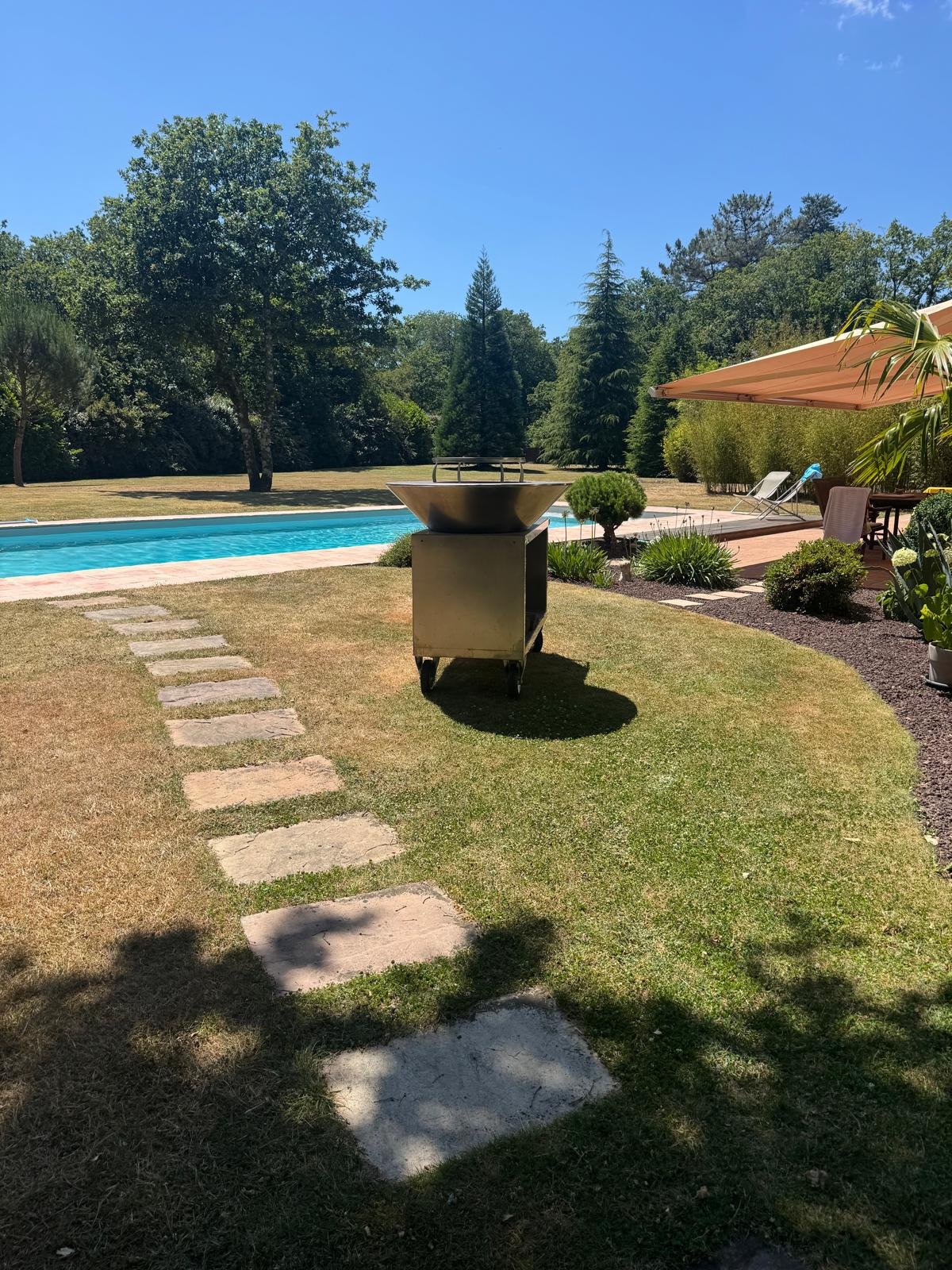 Espace extérieur avec une piscine, un barbecue en métal, un parasol, des chaises longues, un jardin bien entretenu et des arbres en arrière-plan sous un ciel bleu.