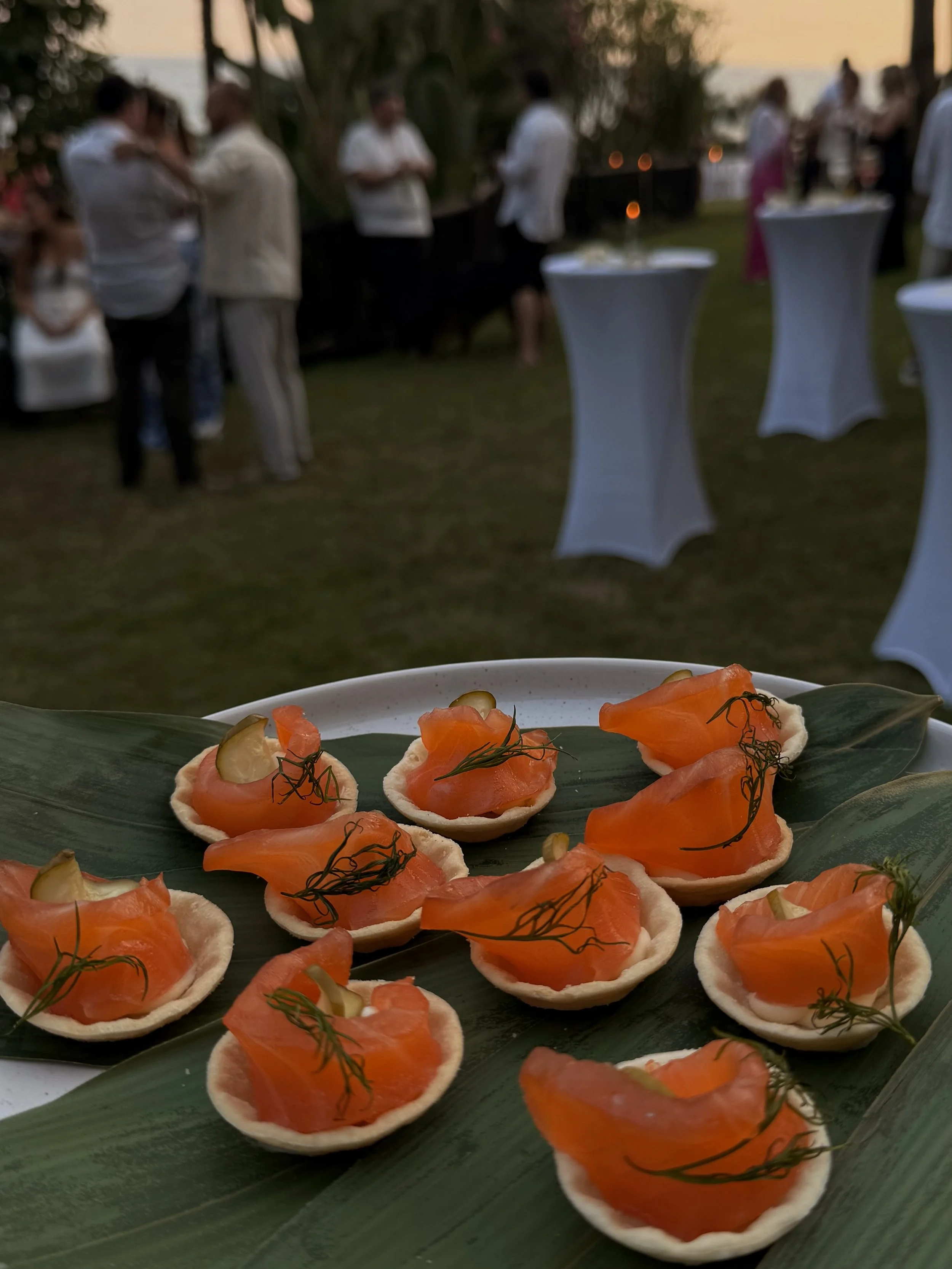 Close-up of salmon canapé appetizers garnished with dill and pickle slices, served on a leaf-lined tray at an outdoor event during sunset with guests and cocktail tables in the background.