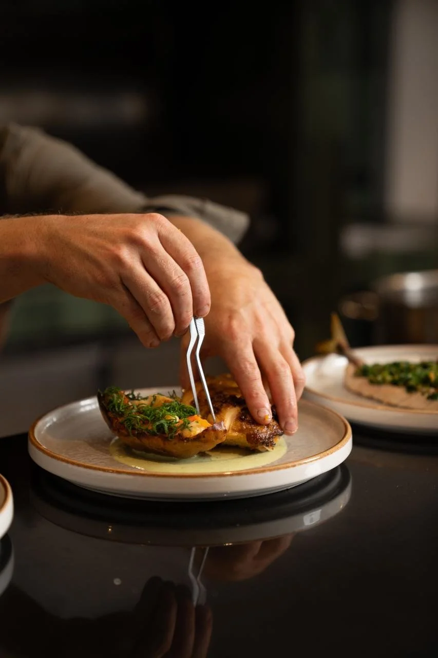 A chef garnishing a cooked fish with herbs on a ceramic plate.