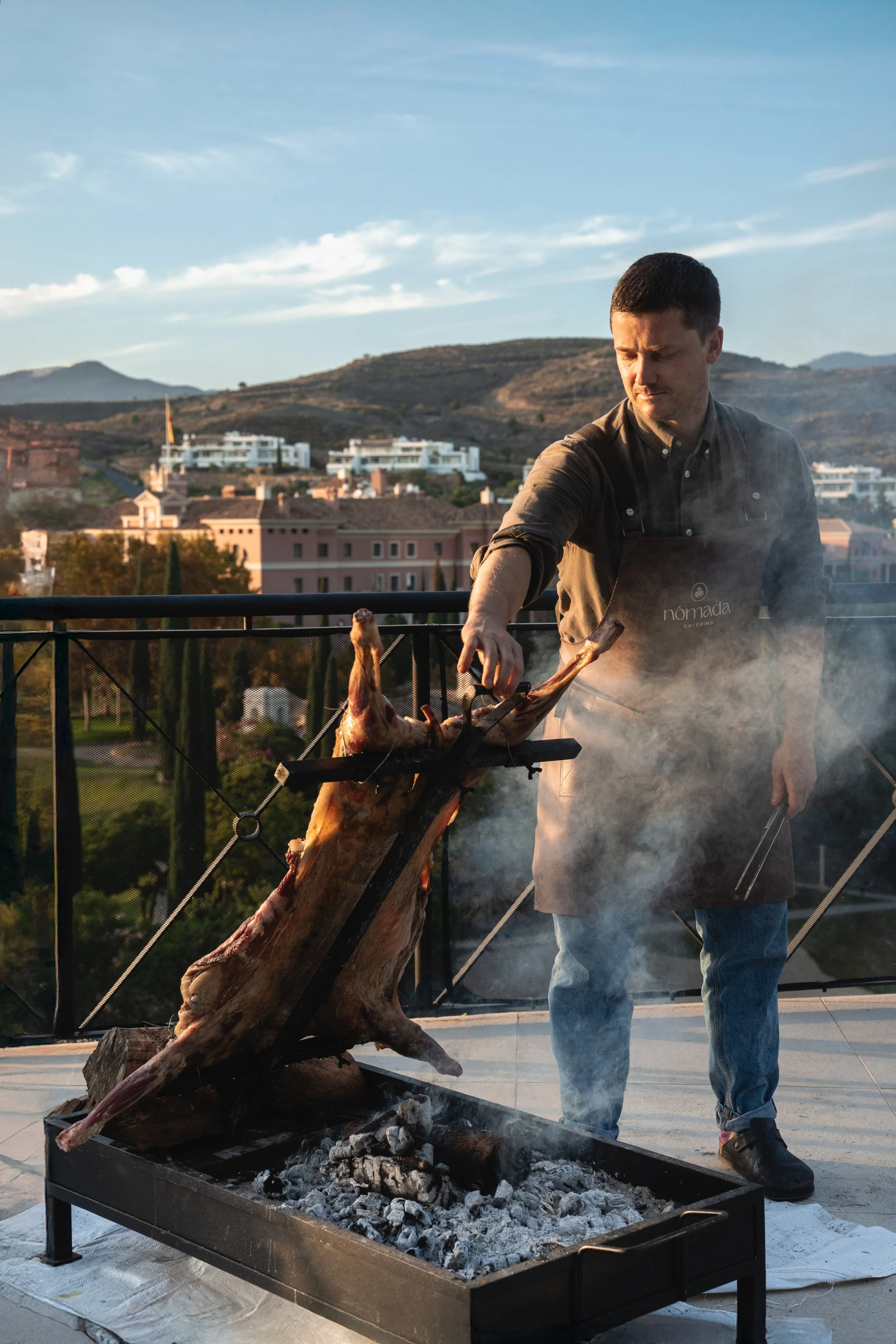 Man roasting a whole animal on an open fire grill outdoors with a mountain landscape in the background.