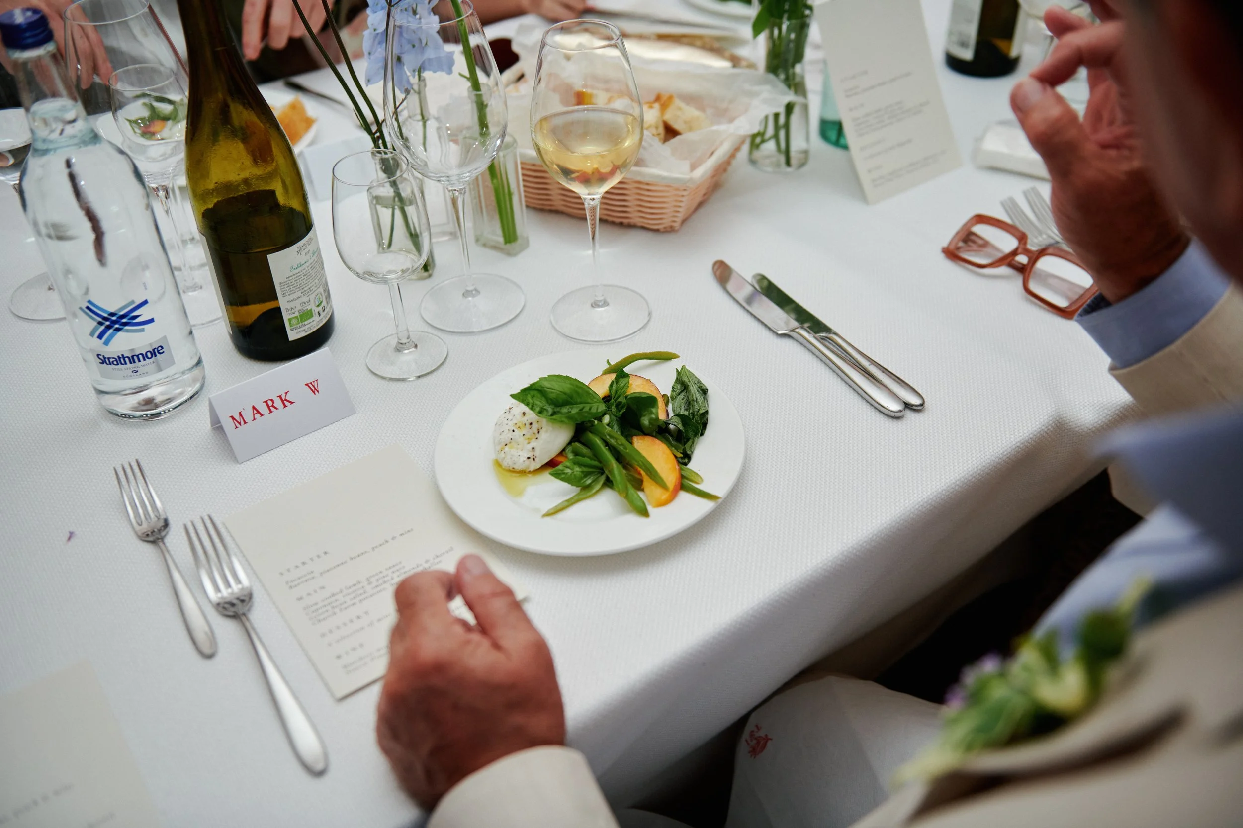 A dining table set with glassware, a wine bottle, a water bottle, a plate of salad, and a person with glasses and a light-colored suit. The table has a name card labeled 'MARK W', and there is a menu or notes on the table.