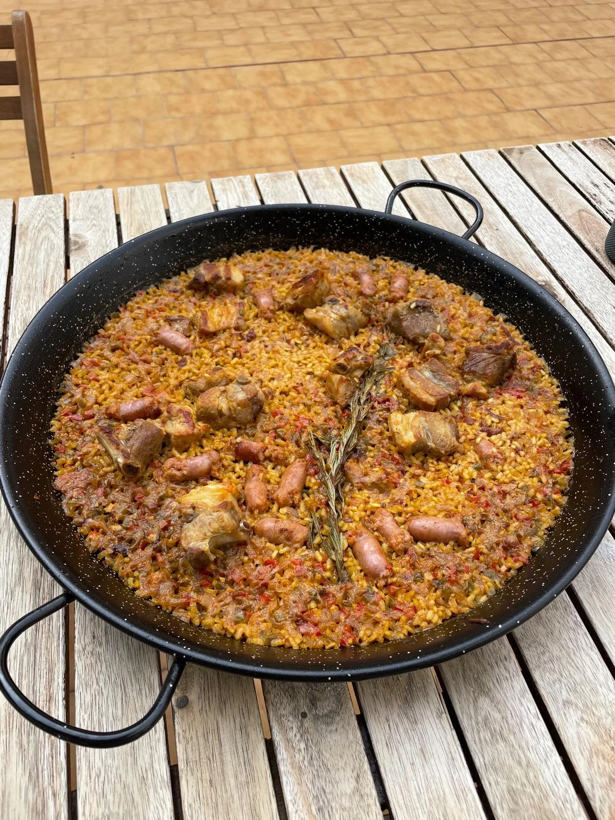 Large paella pan filled with a rice dish containing chicken, sausage, and herbs, garnished with a sprig of rosemary, on a wooden table.