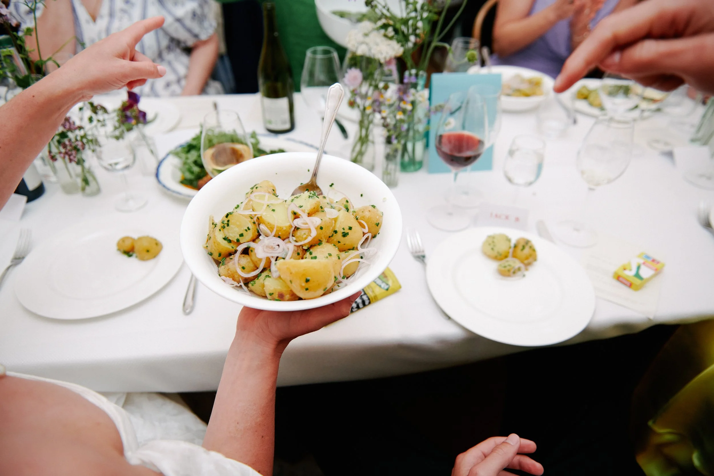 Person holding a bowl of potato salad garnished with green herbs and sliced onions at a dinner party with a white tablecloth, wine glasses, flowers, and other dishes.