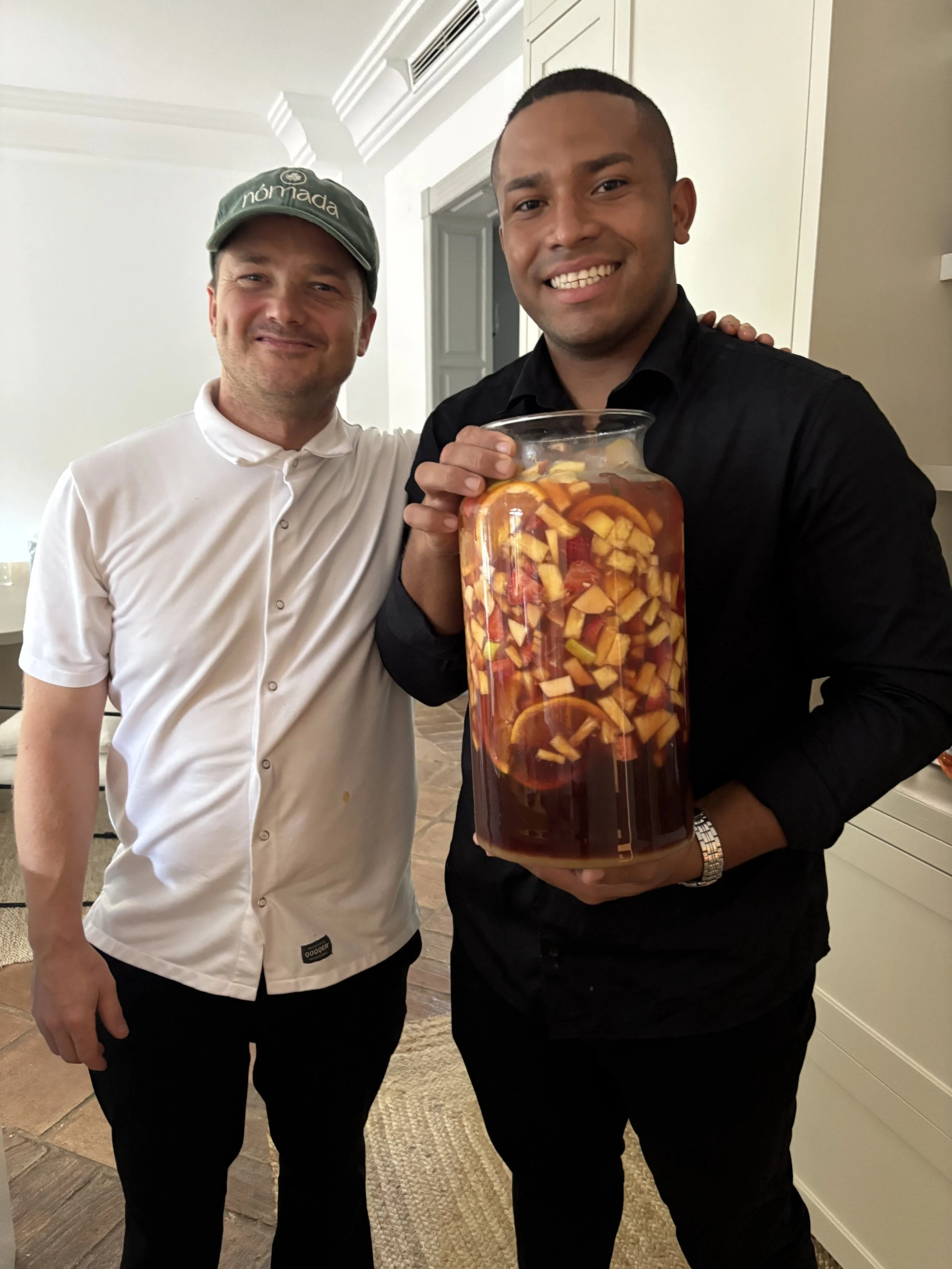 Two men smiling; one holding a large jar with sliced fruit and liquid, others are in a room with white walls and wood flooring.