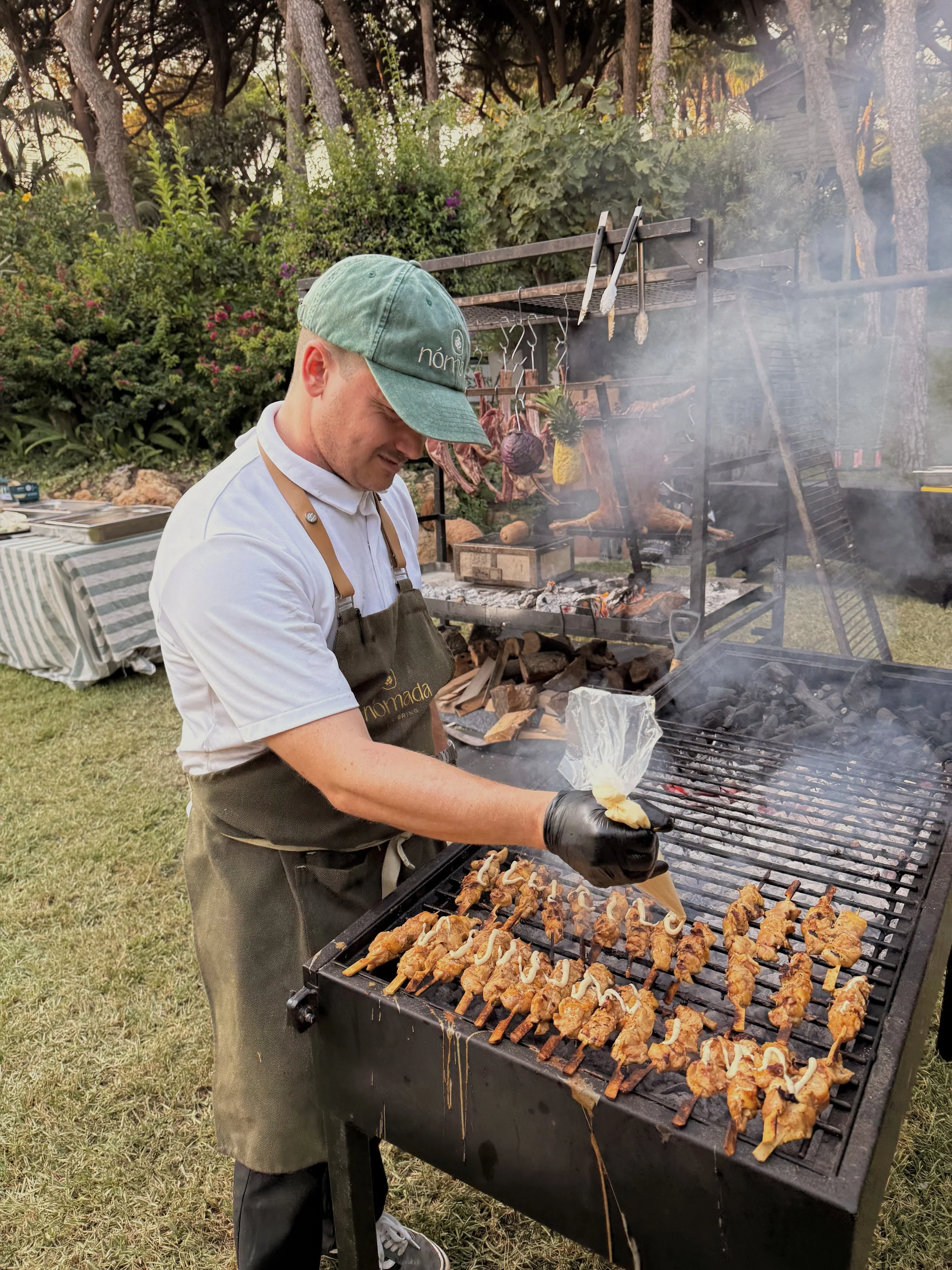 Man grilling skewers of meat and vegetables at an outdoor barbecue, with greenery and trees in the background.