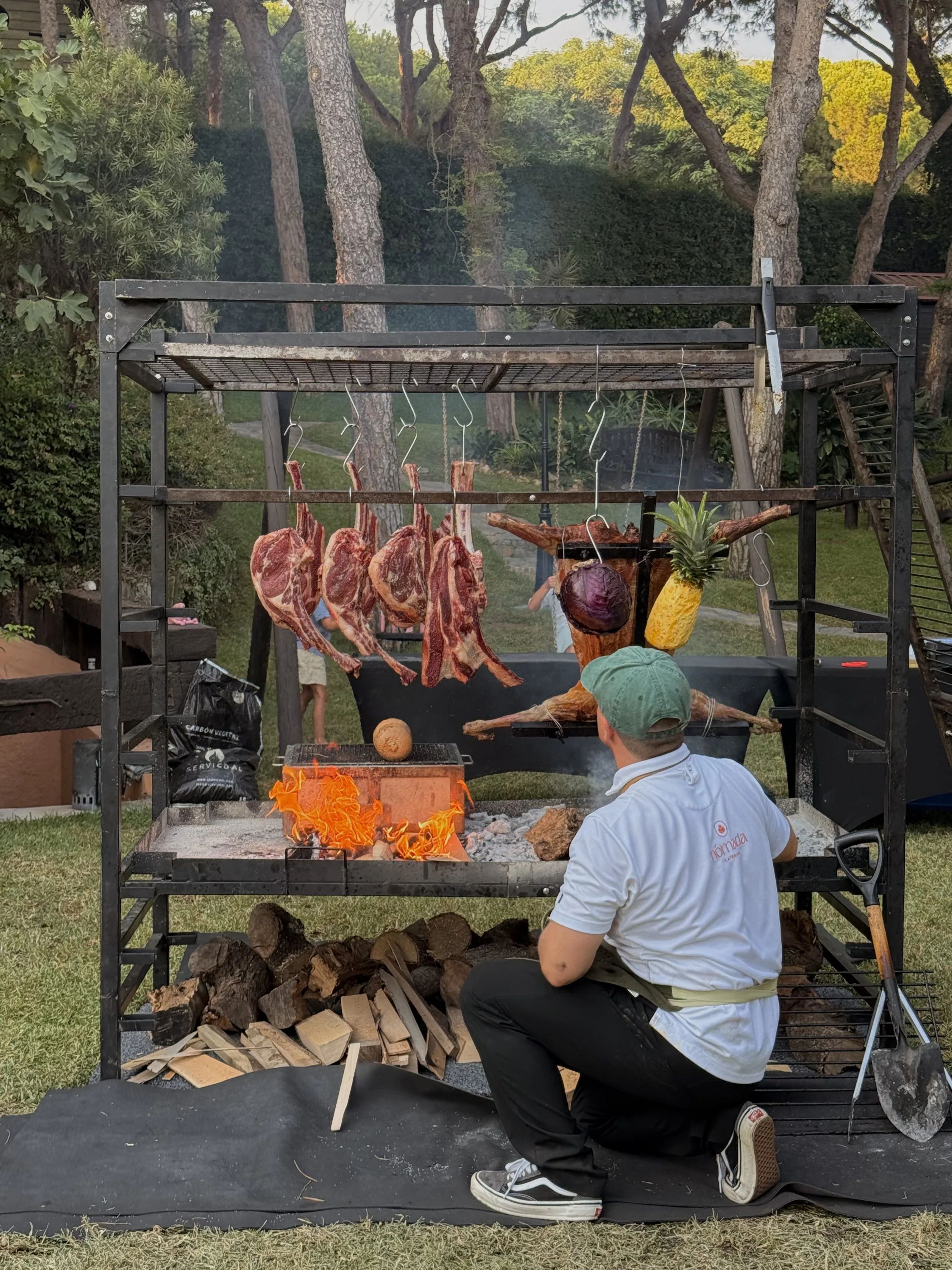 Butchered steaks hanging on hooks over an open fire grill with flames, with a man in a green cap and white apron kneeling in front, preparing to cook outdoor barbecue in a backyard with trees and stacked wood underneath the grill.