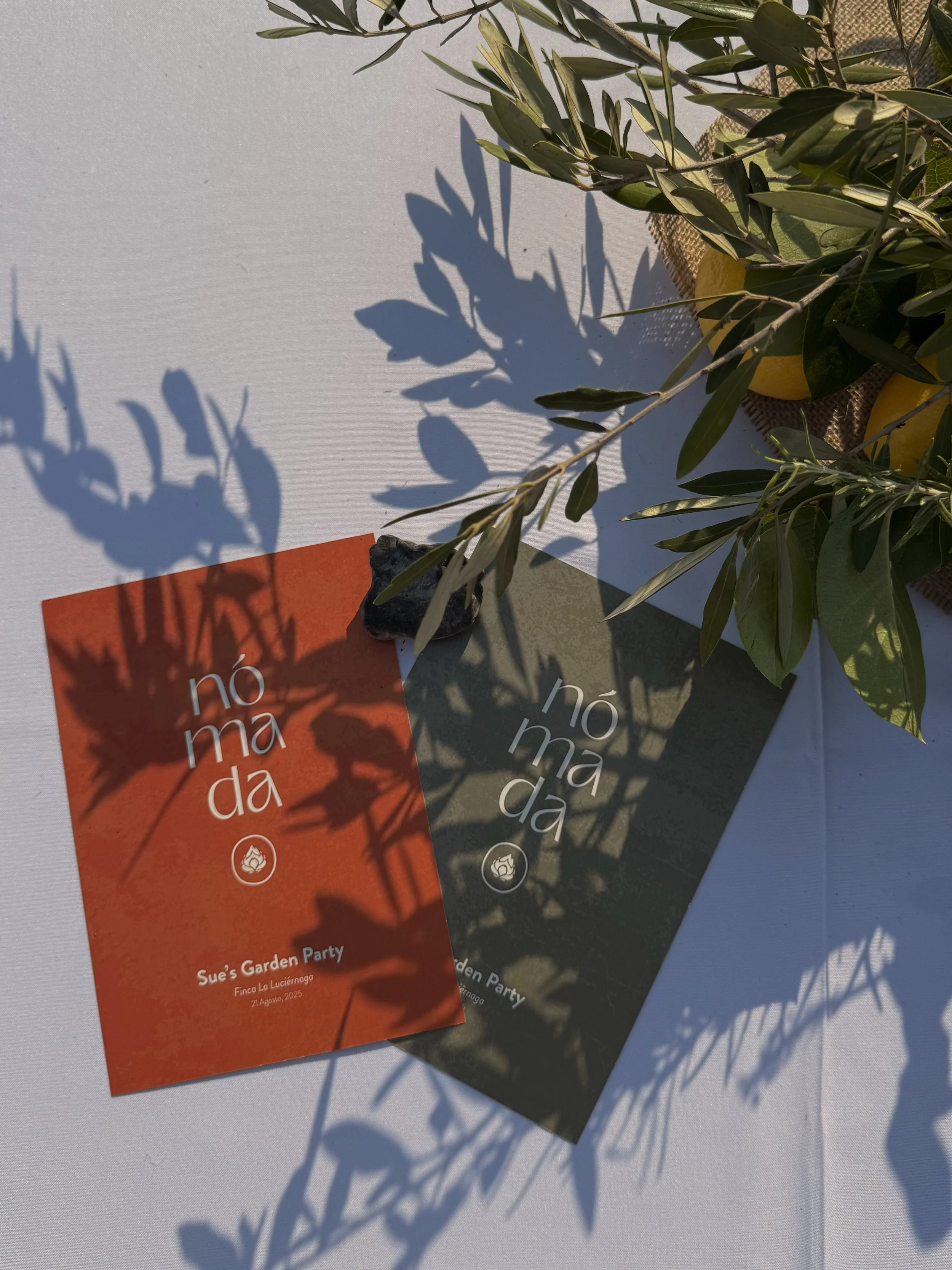 Two event flyers for a garden party, one orange and one dark green, with shadows of leaves cast over them on a white surface, and a branch with green leaves and yellow lemons nearby.