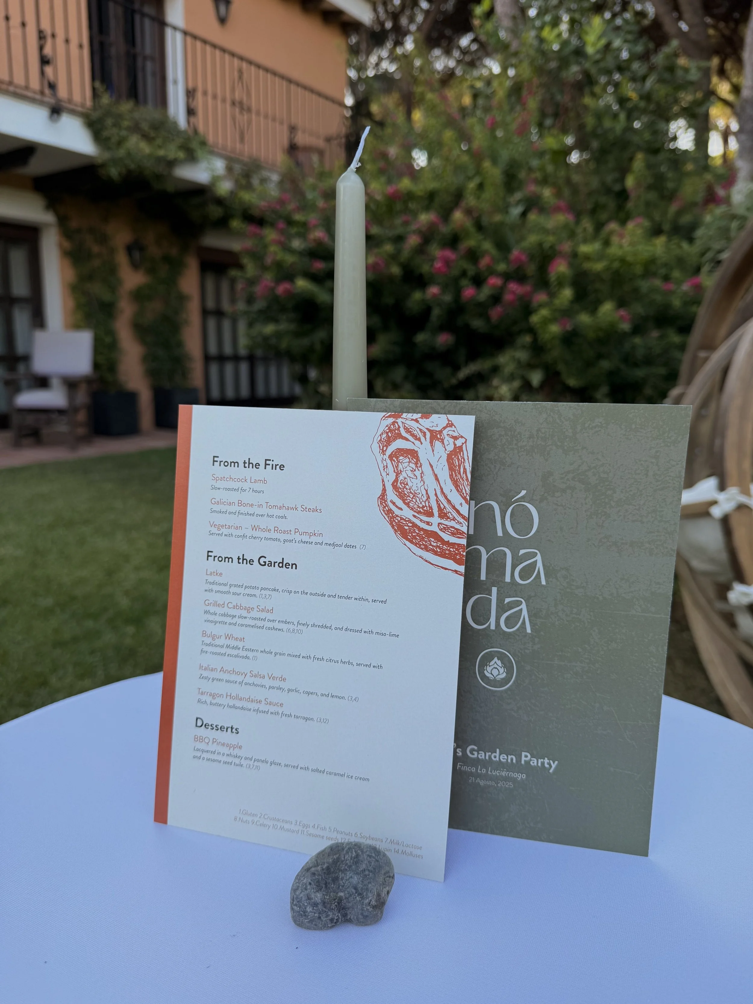 A table with a menu, a candle, and a small rock in an outdoor garden setting with trees and a building in the background.